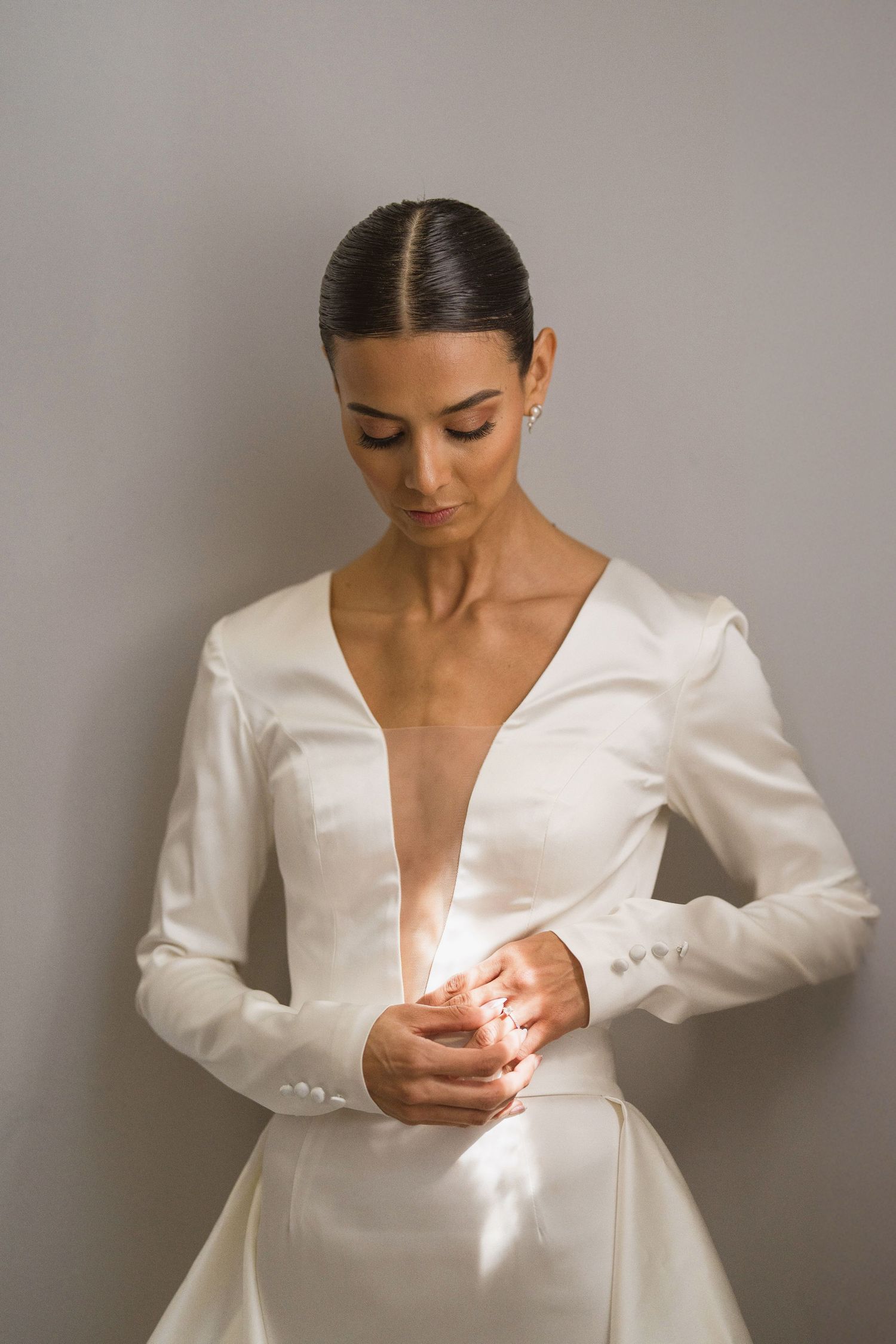 Bride in white satin deep-V wedding dress looking down gracefully against gray wall.