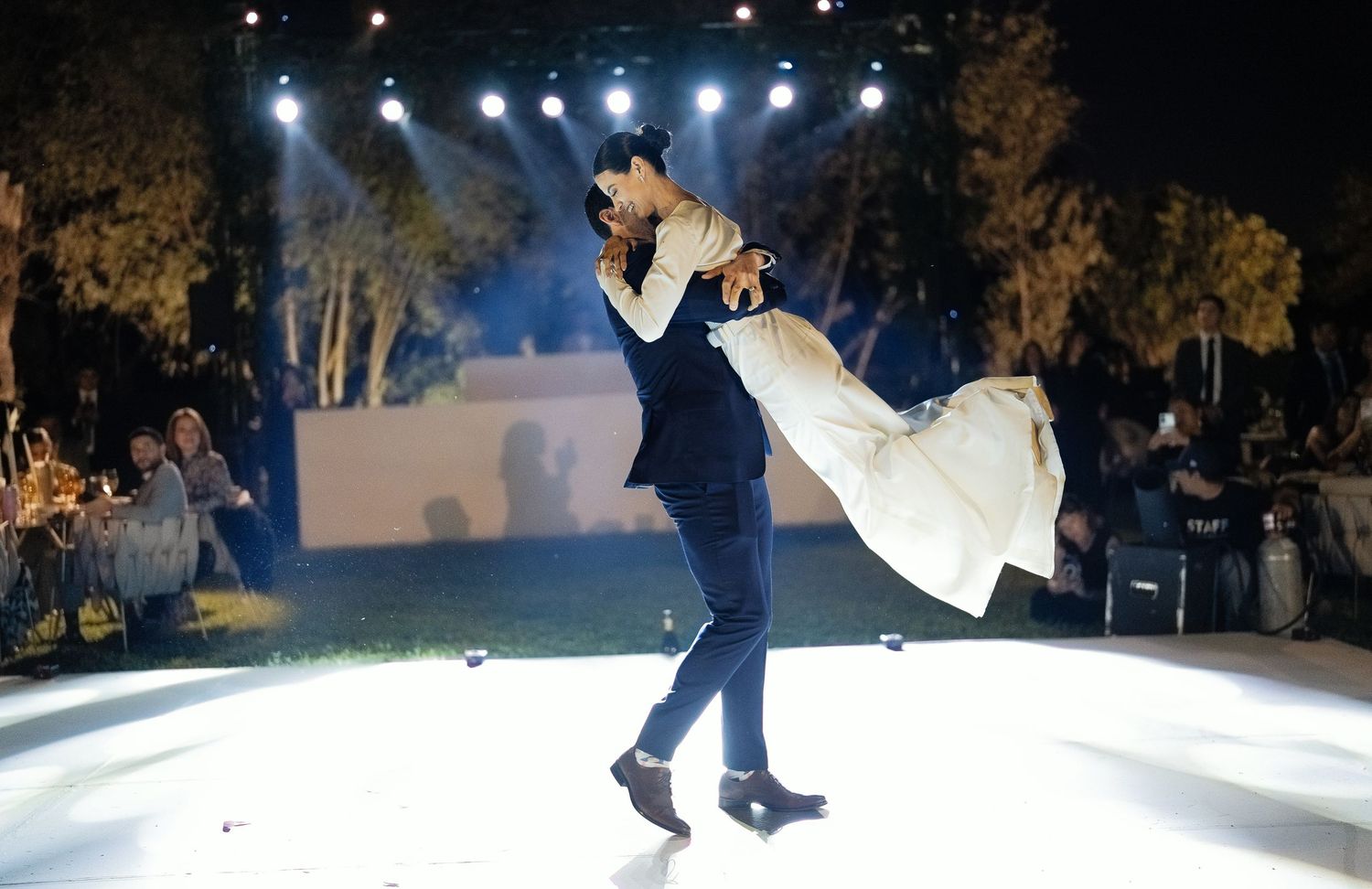 Groom lifting bride in white dress during romantic first dance under bright wedding stage lights.