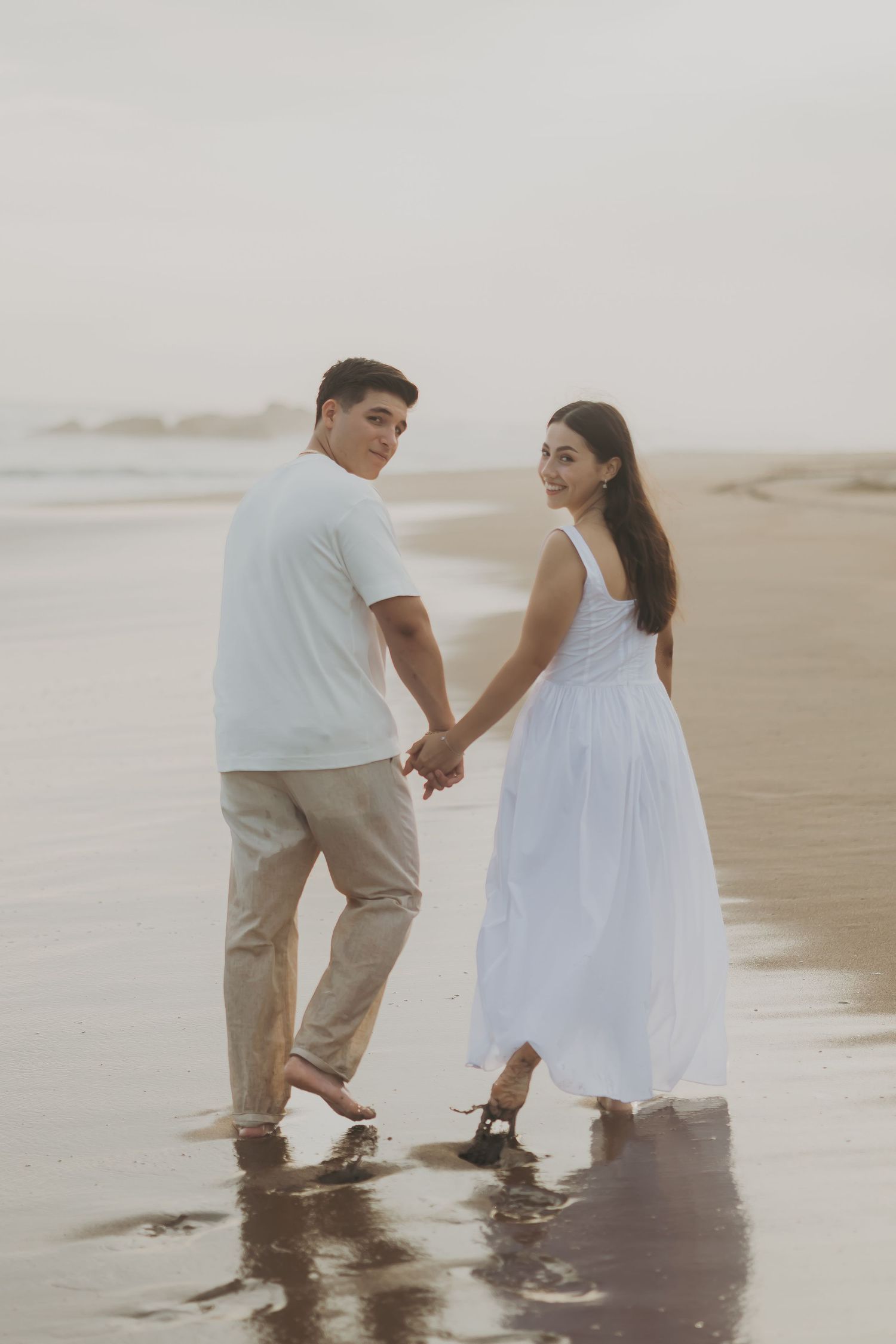 Couple holding hands walking barefoot on a beach, woman in white dress and man in white shirt and khaki pants.