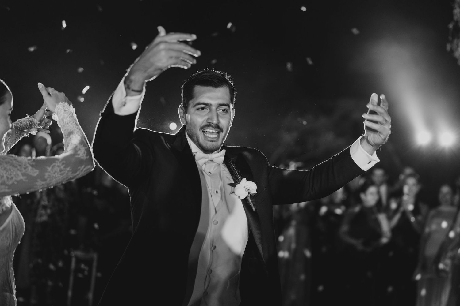 Groom in tuxedo celebrates joyfully with raised arms at wedding reception in black and white photo.