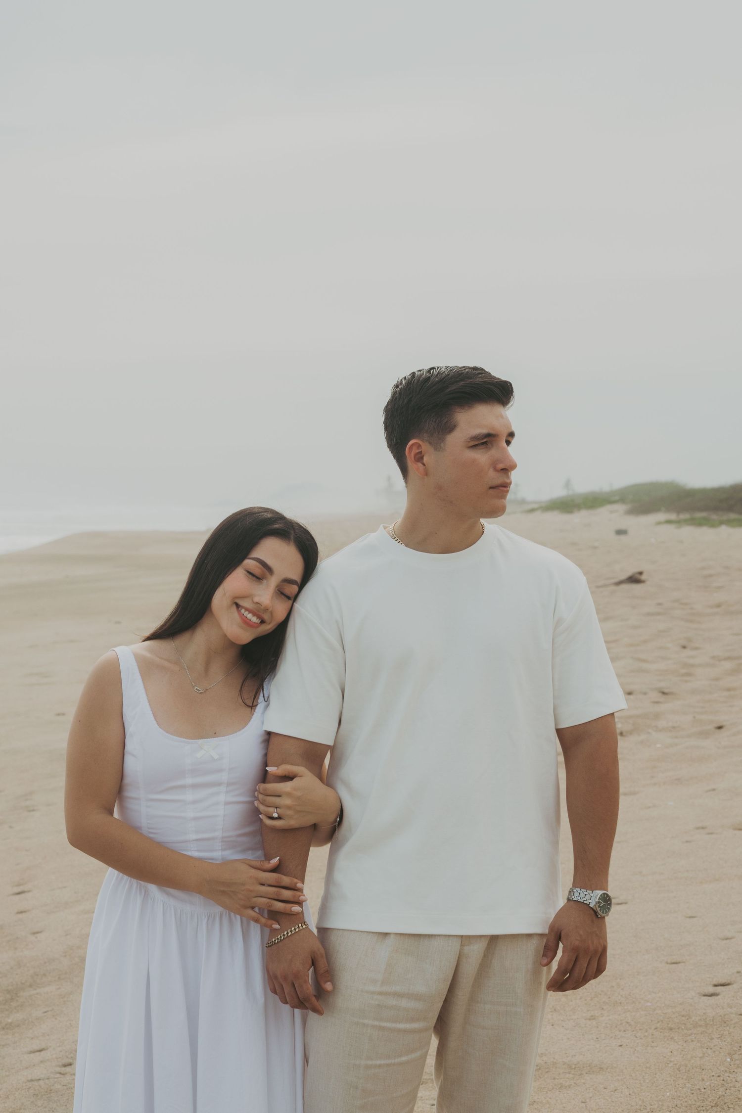 A young couple in white outfits poses on a sandy beach, the woman smiling as she leans against the man.