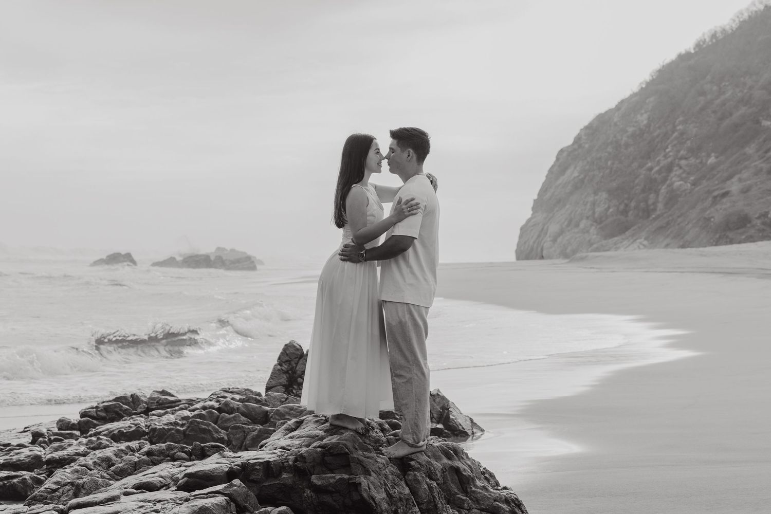 Black and white photo of a romantic couple embracing and kissing on rocky beach with ocean waves and cliffs.