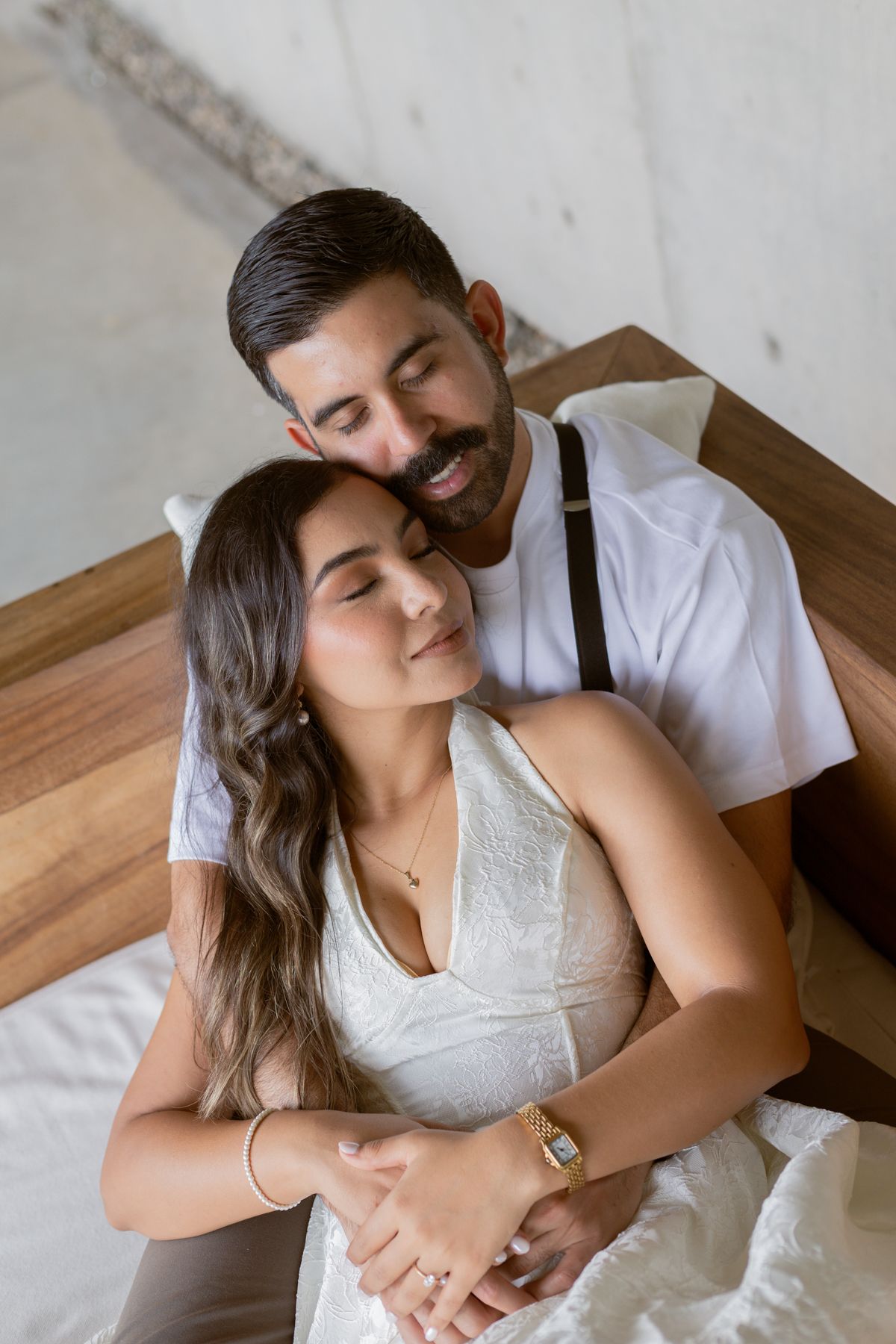 Romantic couple cuddling on a bed, woman in white halter dress with eyes closed, man smiling warmly.