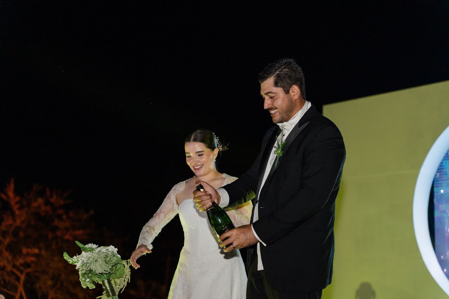 Bride and groom open a champagne bottle together on stage at their outdoor evening wedding reception.