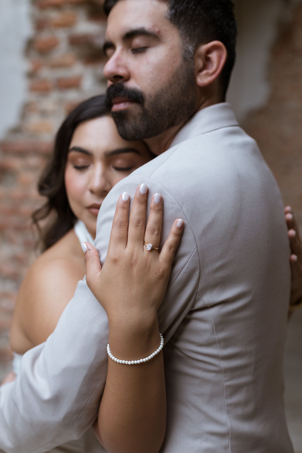 Close-up of bride resting head on groom shoulder, engagement ring and pearl bracelet visible.