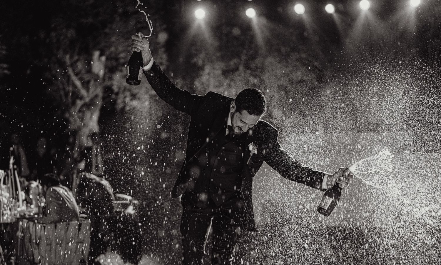 Groom sprays champagne bottles in dramatic black and white wedding celebration photo under stage lights.