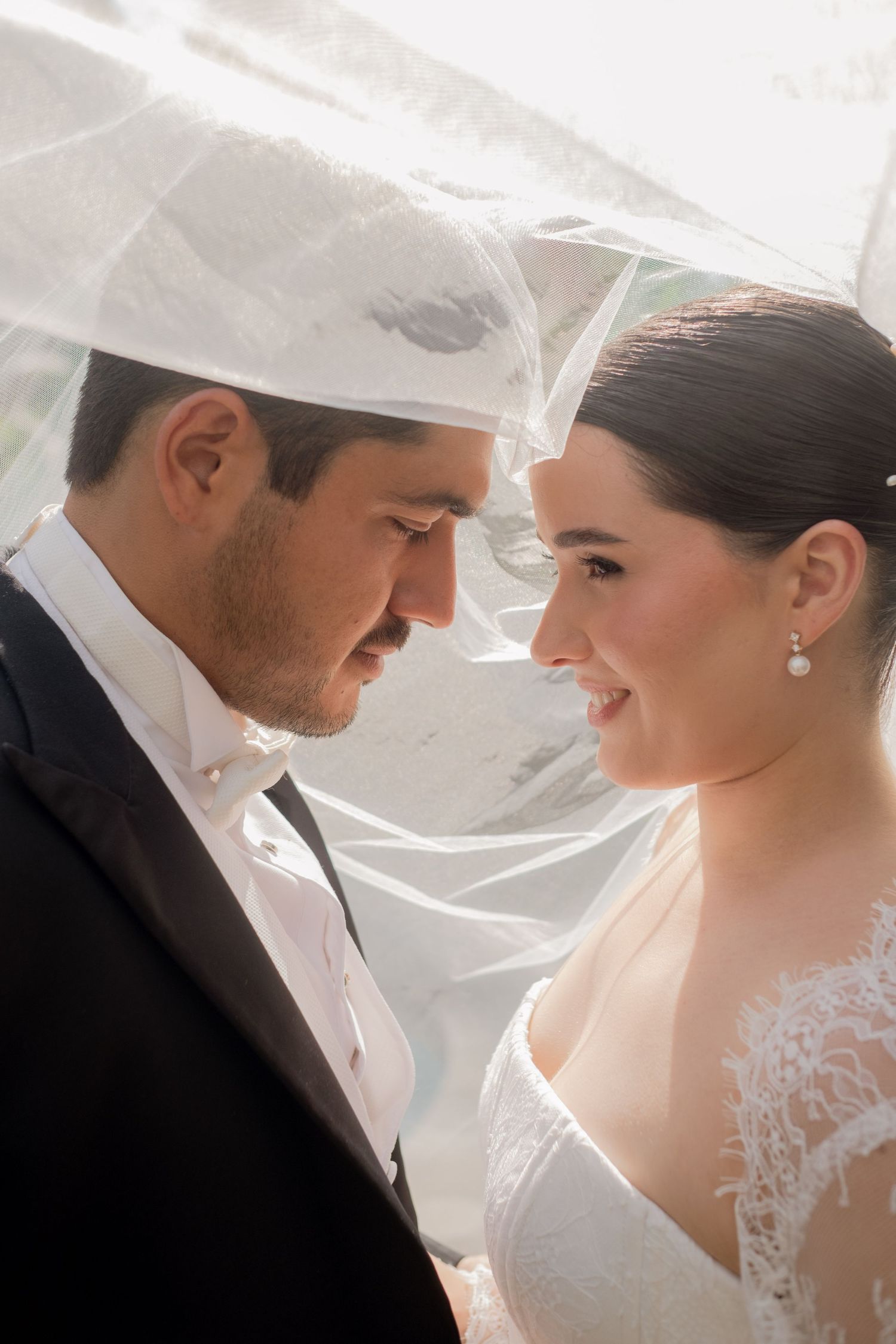 Smiling bride and groom gazing at each other under a white veil in warm sunlight.