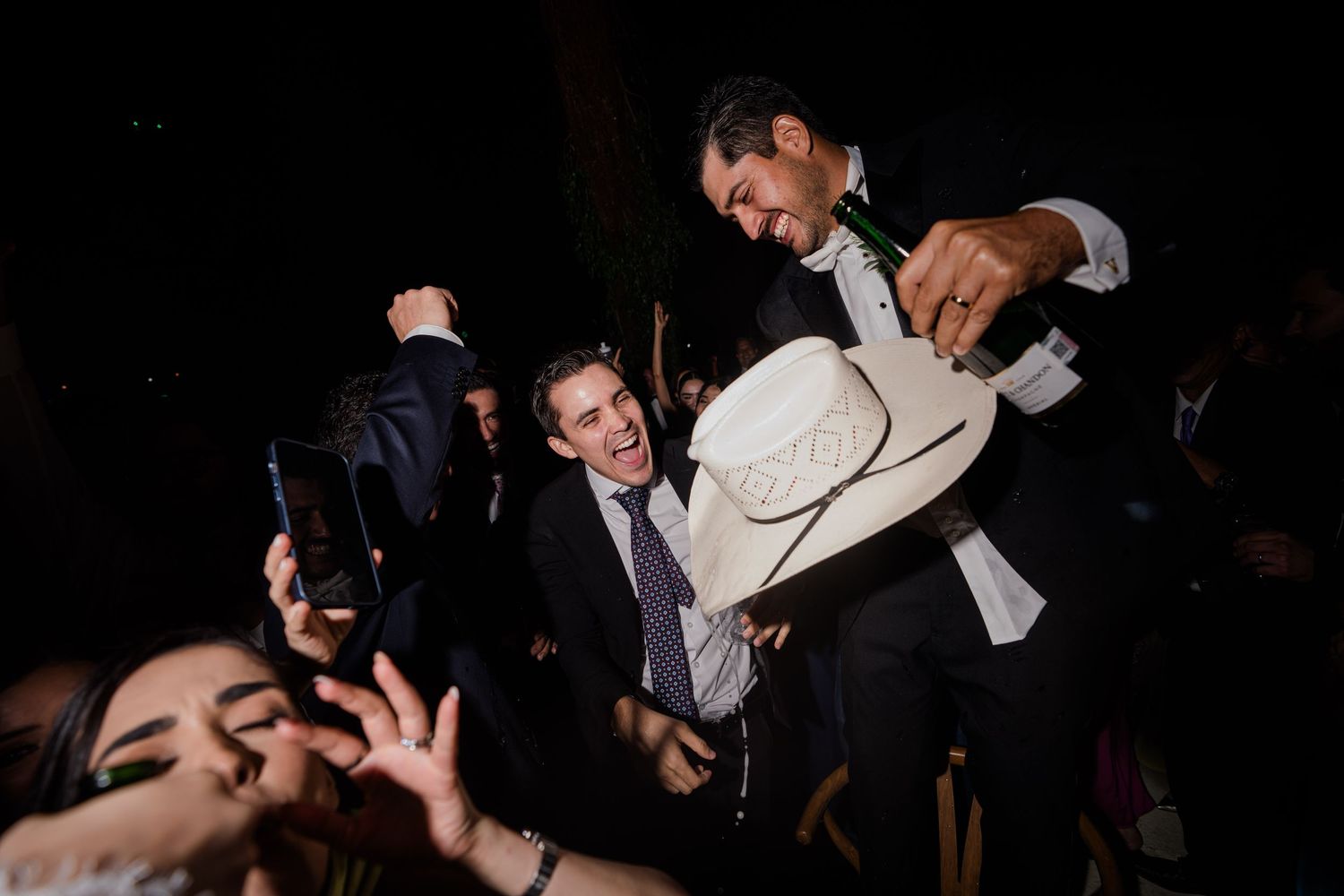 Groom holding white cowboy hat above crowd of excited guests at wedding reception.