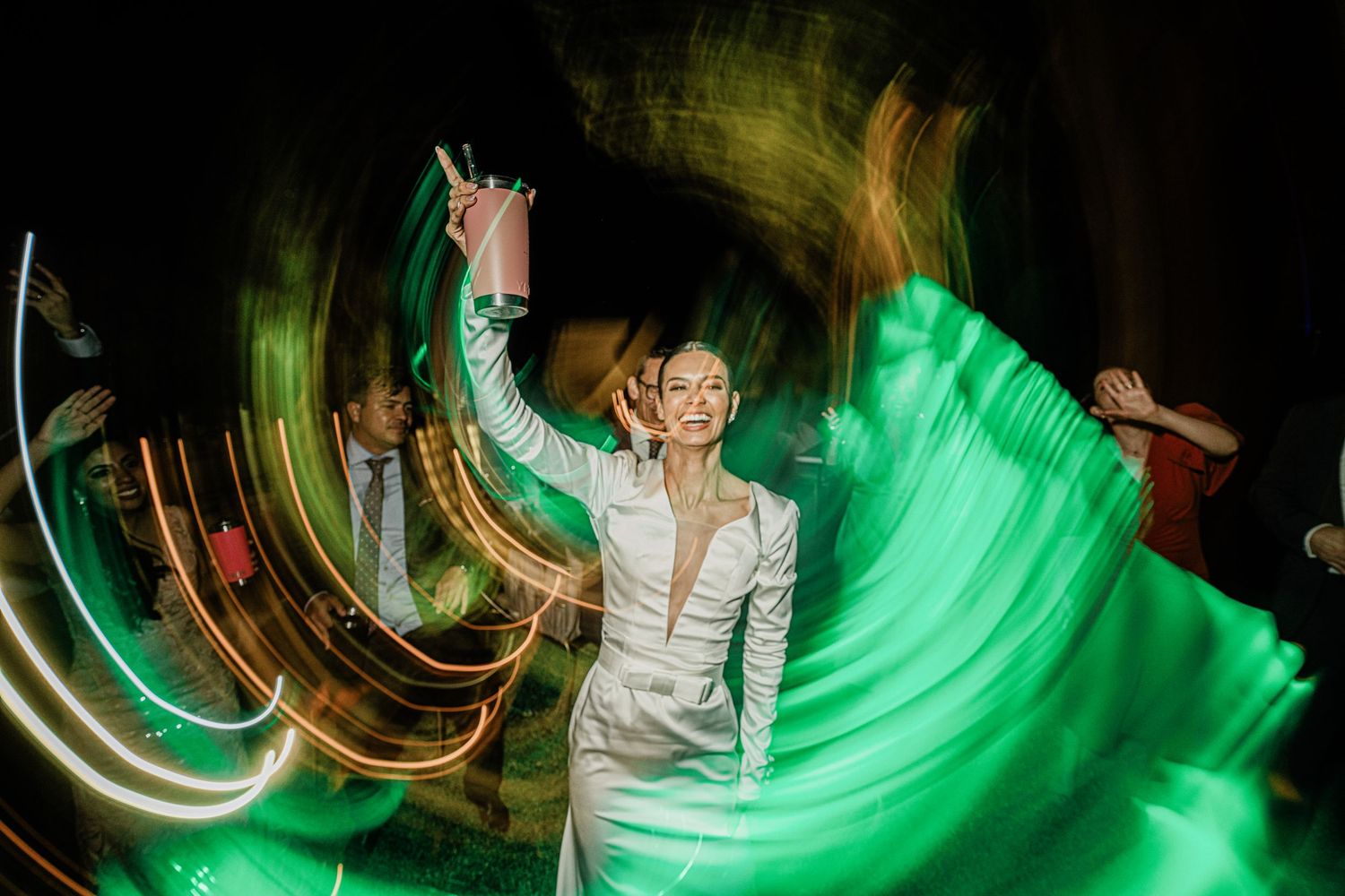Bride in fitted white gown dances with arms raised surrounded by colorful light trails at reception.