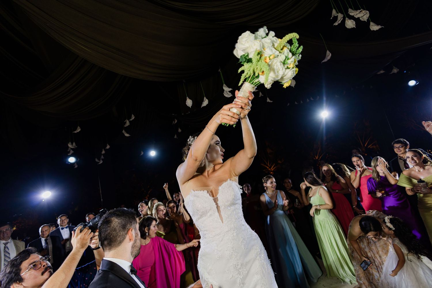 Bride tossing her white floral bouquet to eager guests during a lively wedding reception celebration.