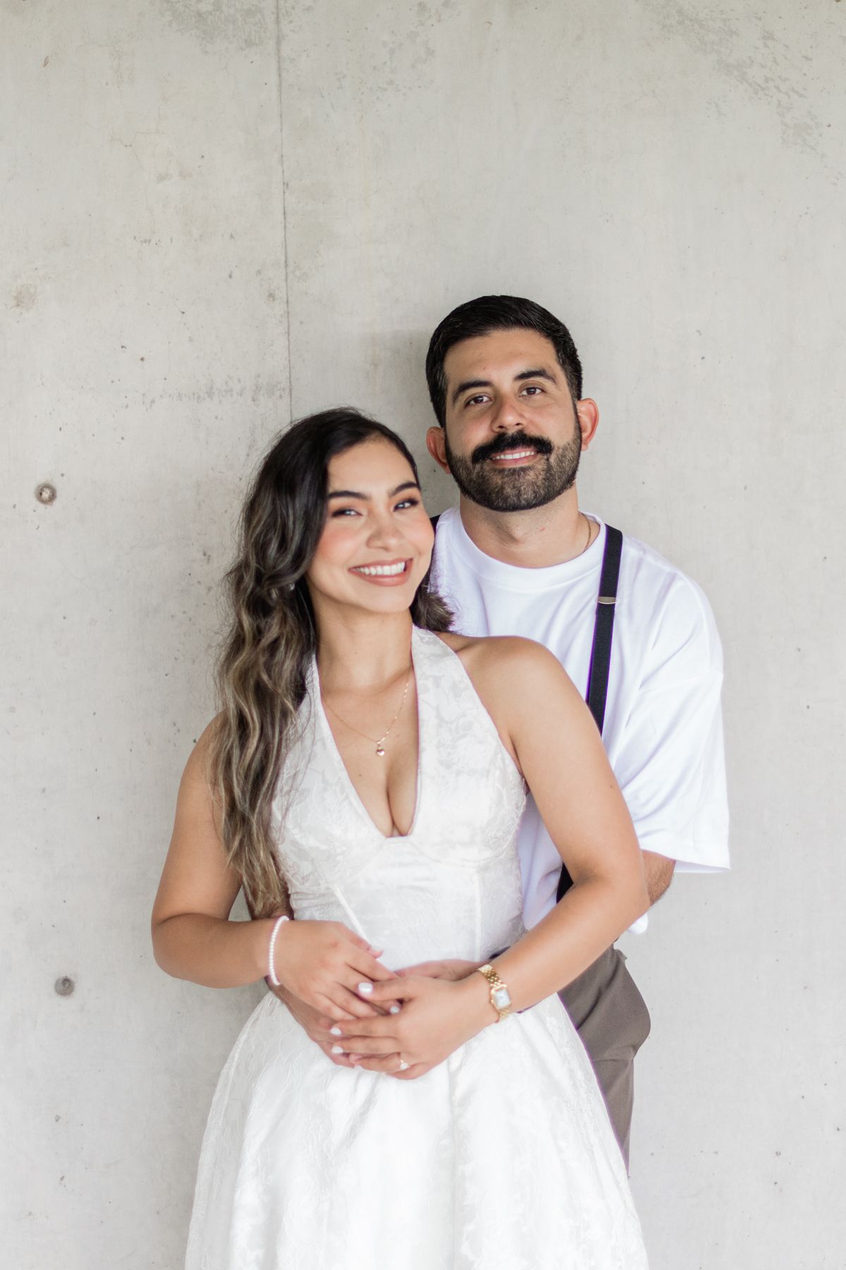 Smiling couple embracing against a concrete wall, bride in white dress and groom in white tee with suspenders.