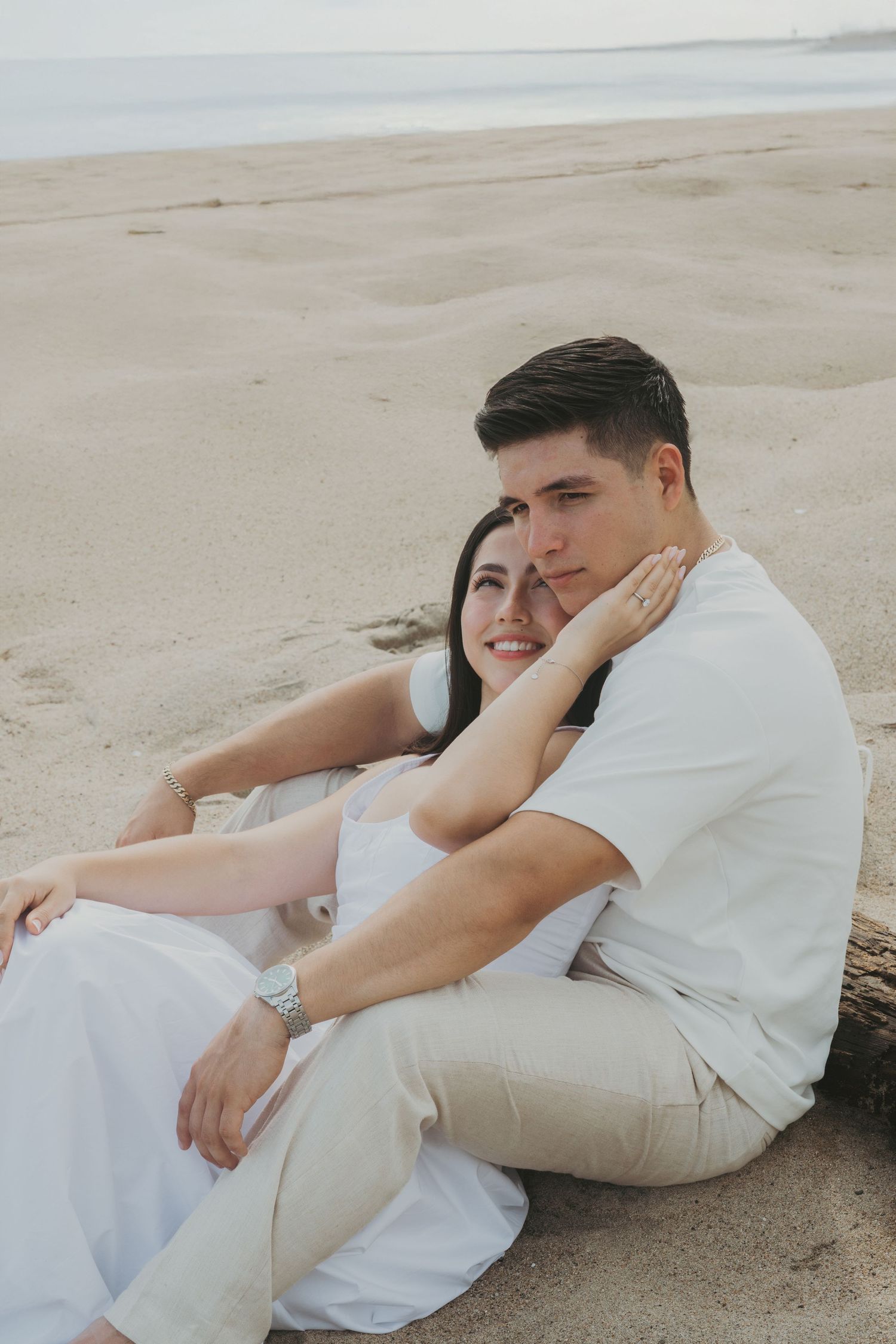 Couple in white outfits sitting together on sandy beach, woman leaning on mans shoulder, serene ocean backdrop.