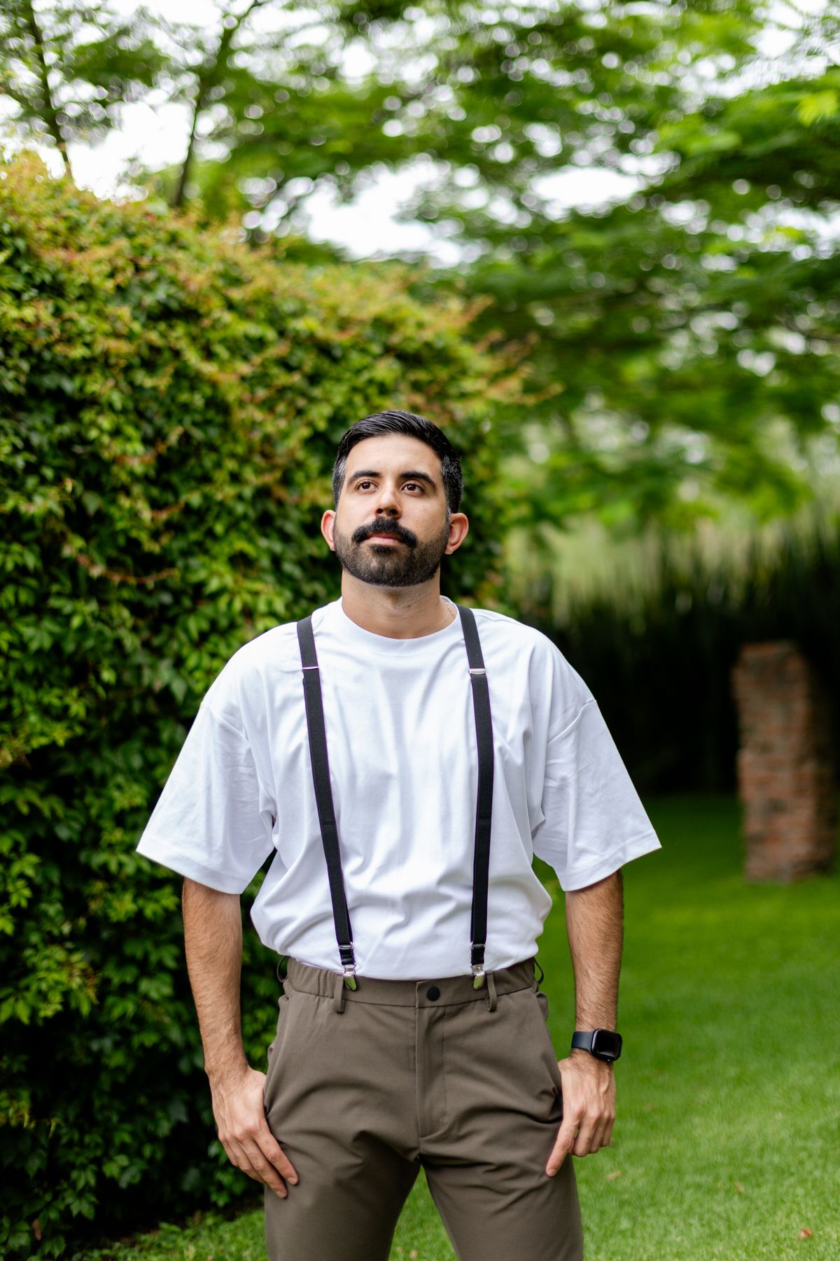 Bearded man in white t-shirt and suspenders stands confidently in a green garden setting.