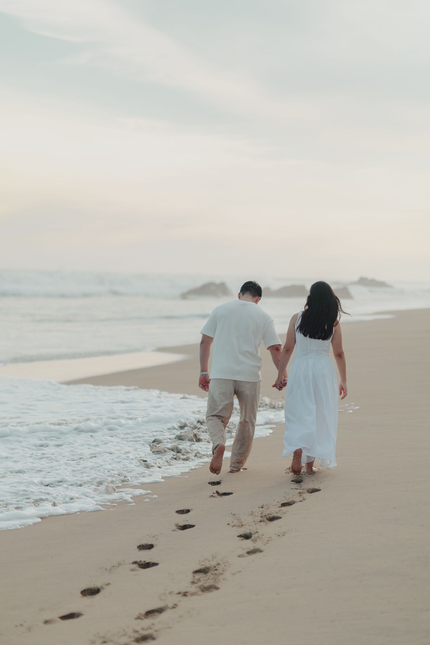 Couple holding hands walking along ocean shoreline leaving footprints in sand at dusk.
