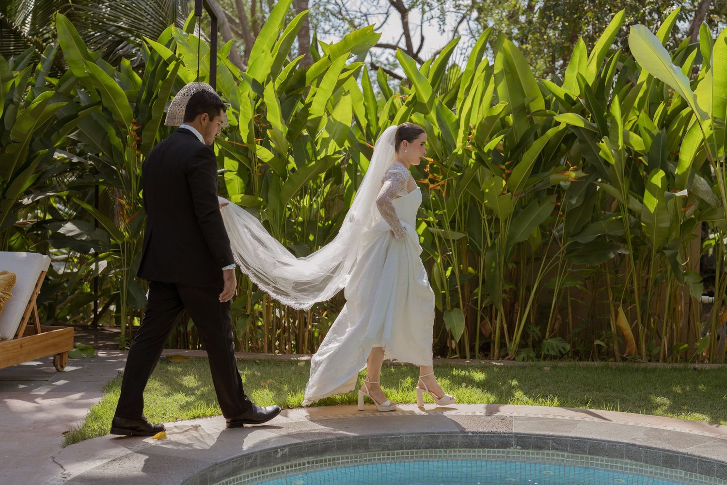 Bride and groom walking hand in hand beside a pool surrounded by lush tropical greenery on their wedding day.