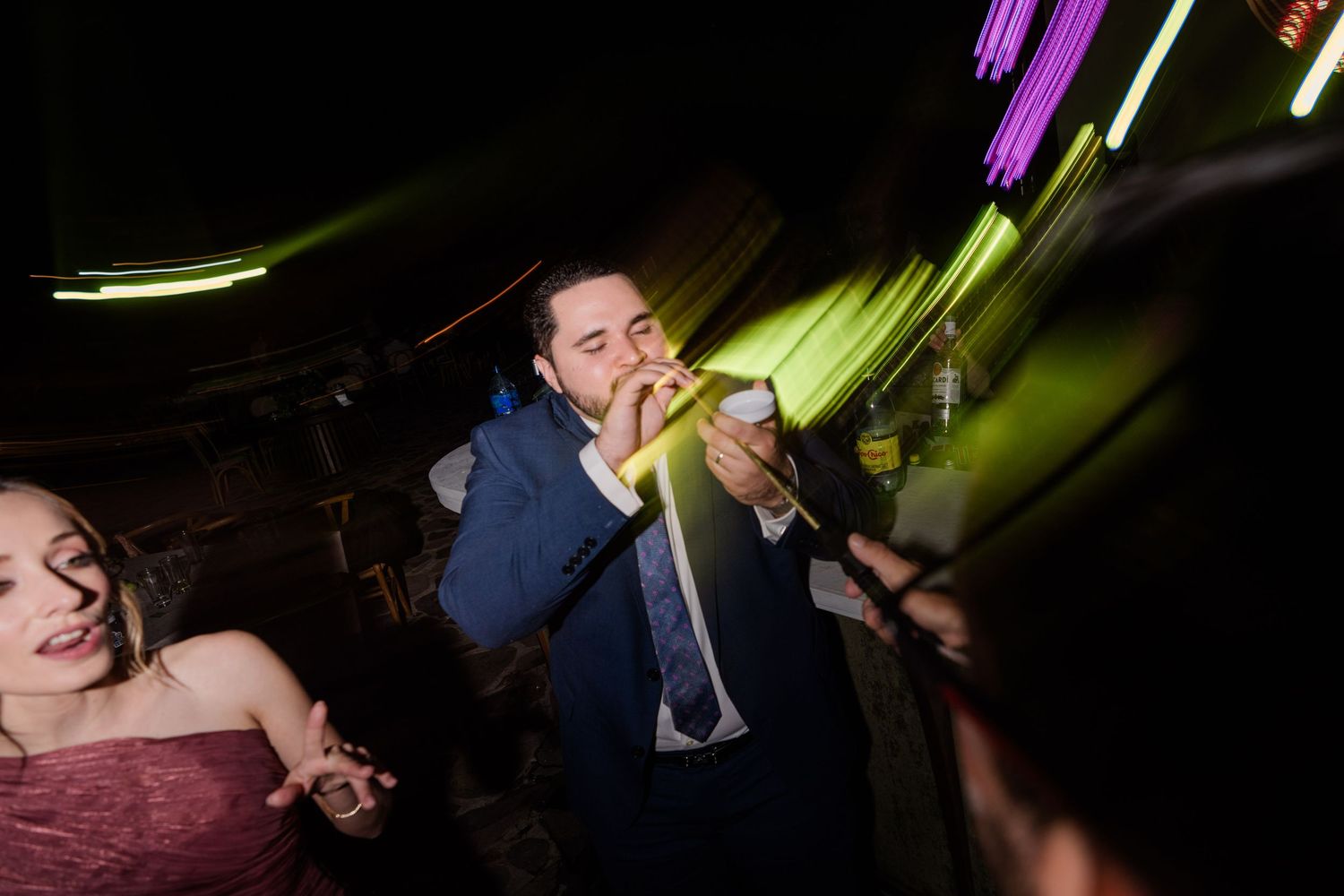 Man in blue suit singing into microphone at lively wedding reception with neon lights.