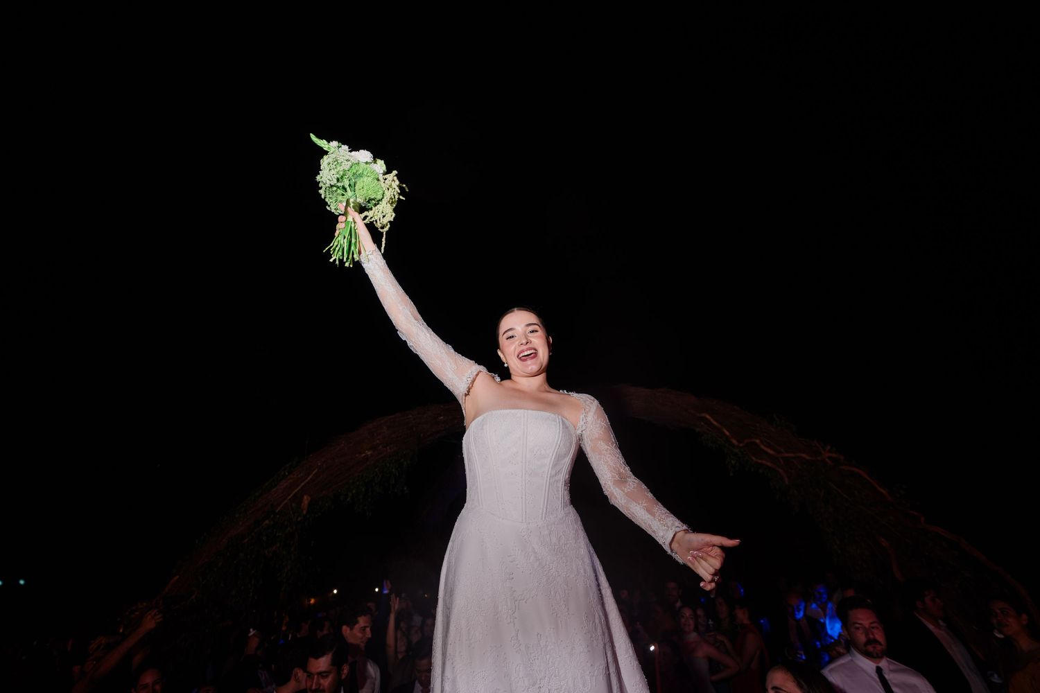 Radiant bride in white gown triumphantly raises her green bouquet overhead at an outdoor nighttime wedding celebration.