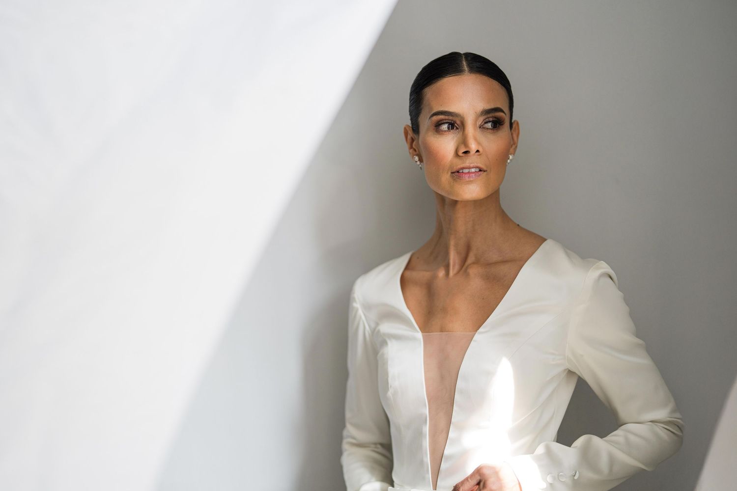 Woman in white deep-V blouse posing elegantly against a light gray wall with dramatic shadows.