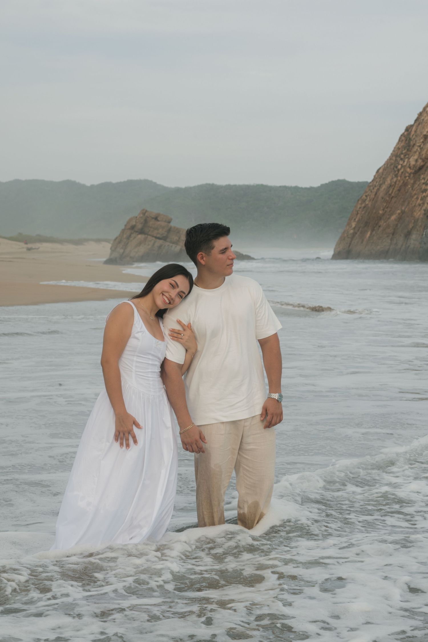Couple in white outfits standing in ocean waves on a sandy beach with rocky cliffs in the background.
