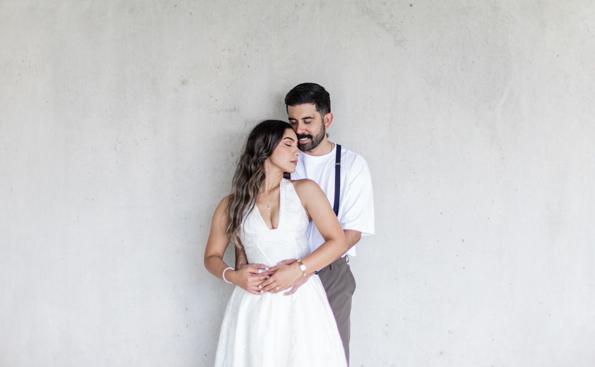 A couple embraces against a light concrete wall, groom in suspenders, bride in white dress.