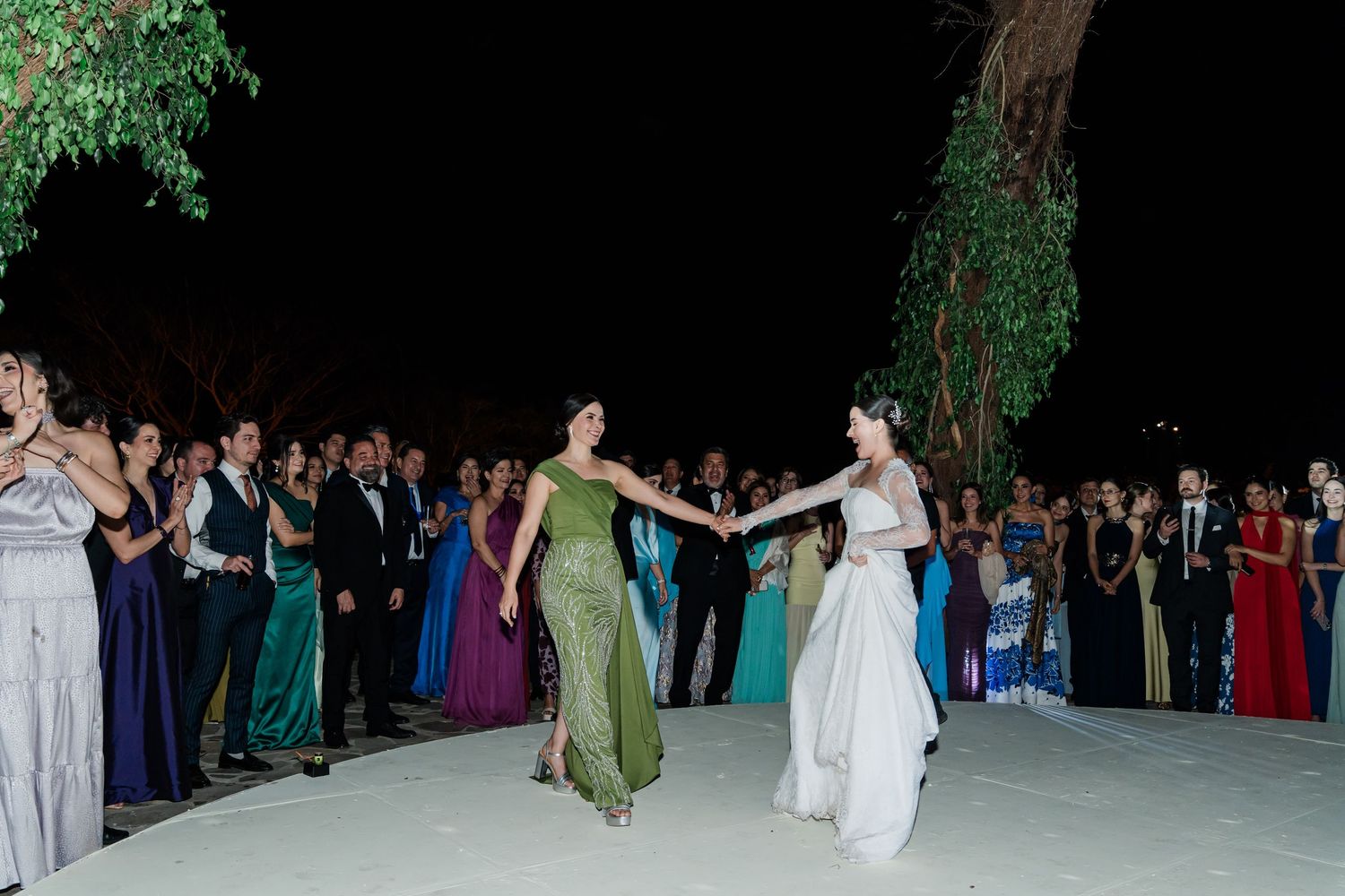 Bride dancing with guest in green gown on outdoor dance floor surrounded by wedding guests.