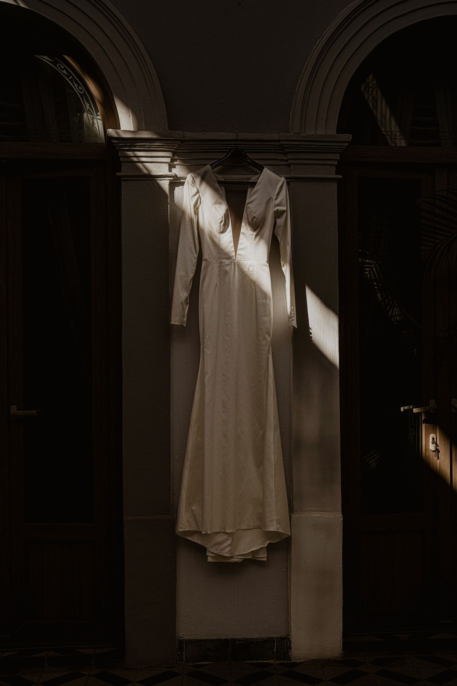 White long-sleeve wedding dress hanging on an architectural column in dramatic moody light.