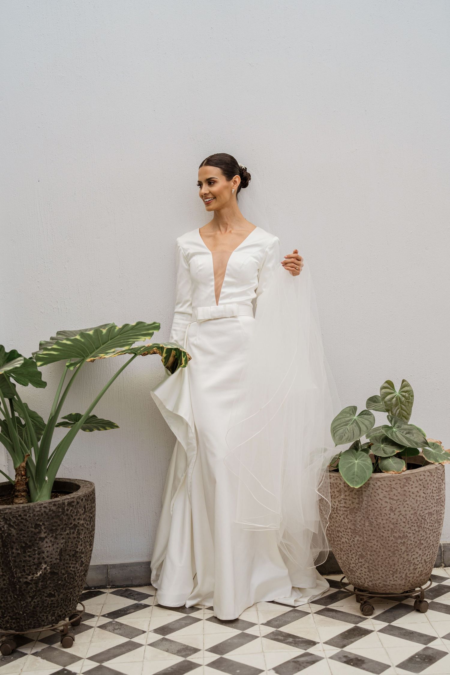 Smiling bride in white V-neck long-sleeve wedding dress holding veil near potted plants.