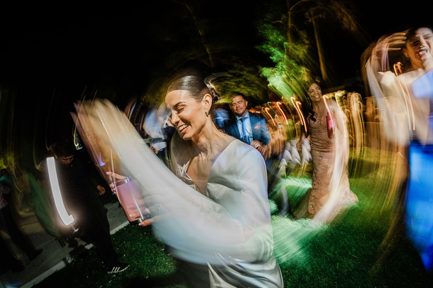 Bride in white gown dancing joyfully with motion blur effect at outdoor wedding reception.
