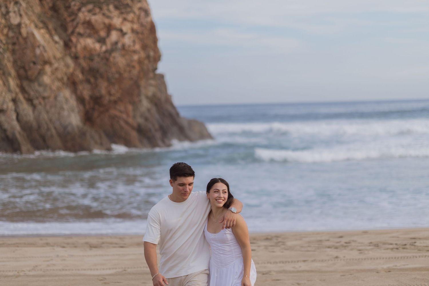 A happy couple walks hand in hand on a sandy beach with rocky cliffs and ocean waves in the background.