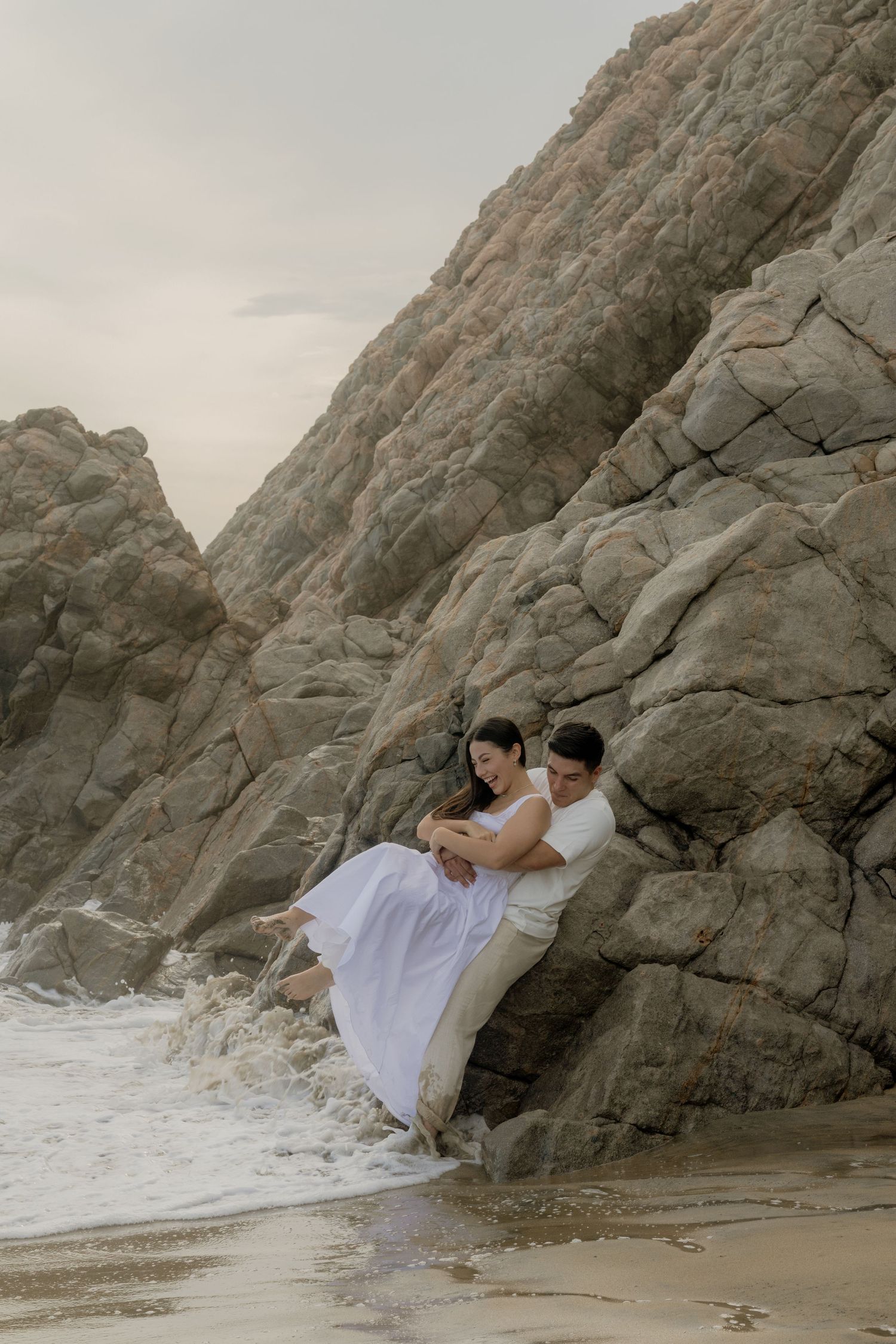 Couple in white sitting against rocky cliffs on a beach, waves washing around them at dusk.