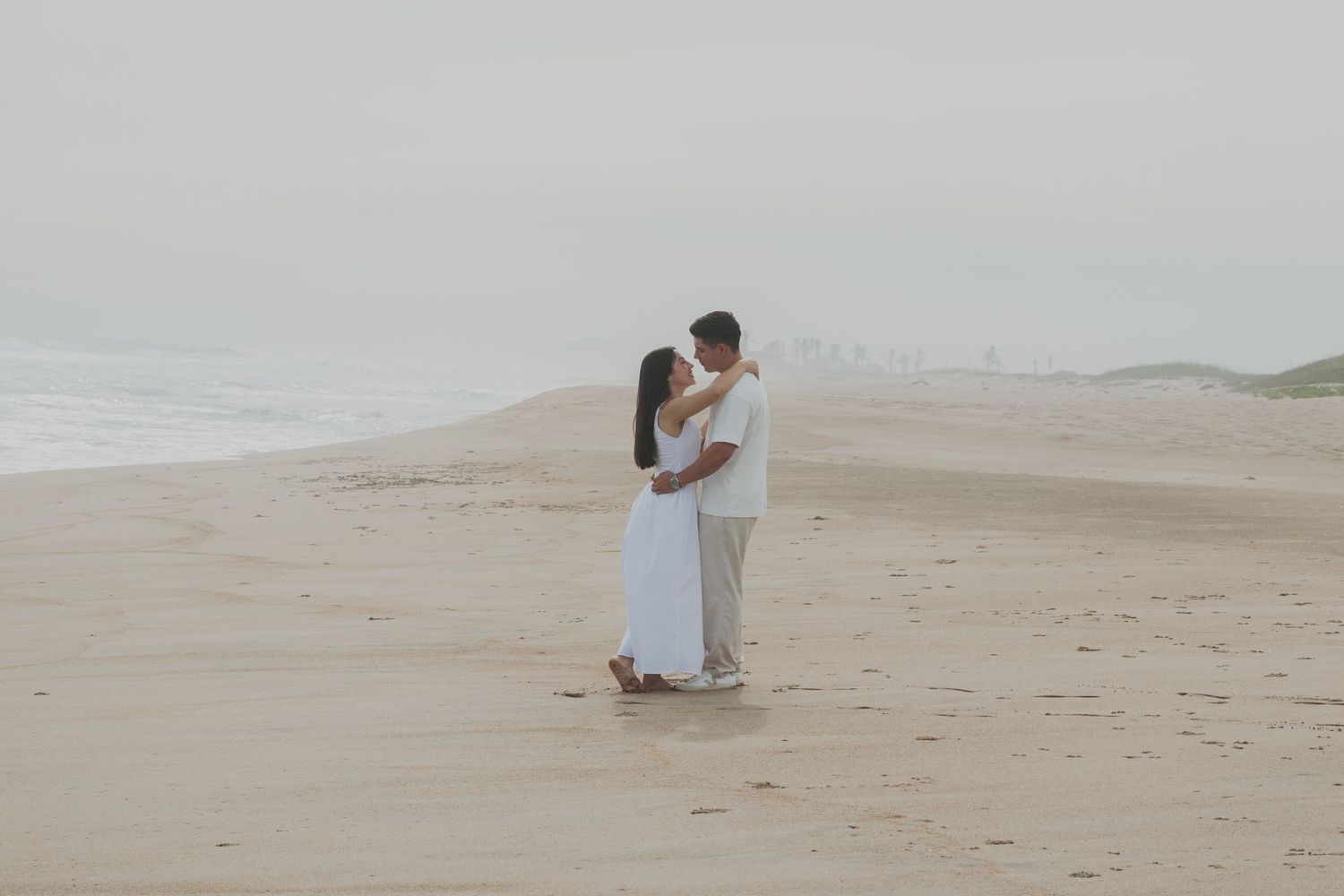 Couple embracing romantically on a misty beach, both dressed in white, with soft sand and ocean waves in the background.