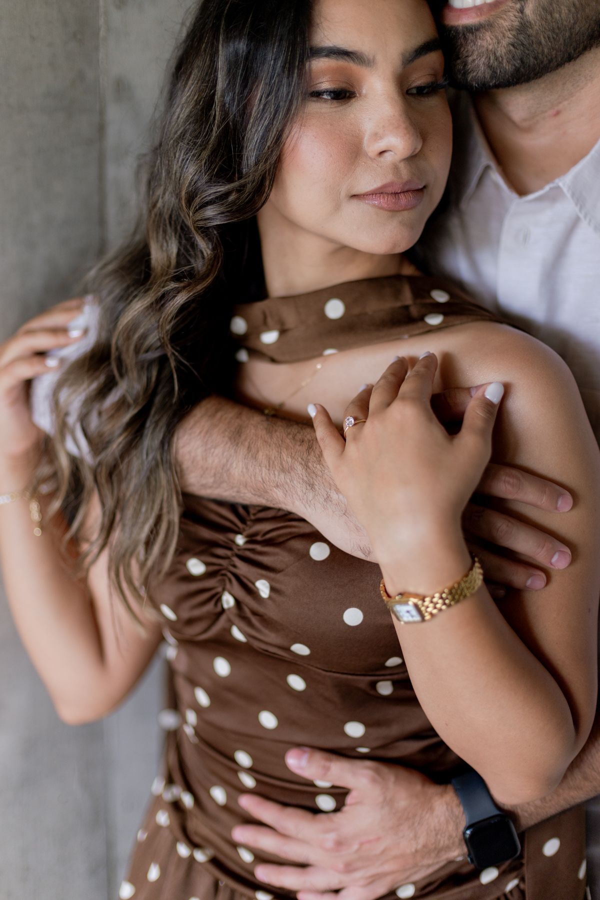 Close-up of couple embracing, man holding woman in brown polka dot dress, gold watch visible on wrist.