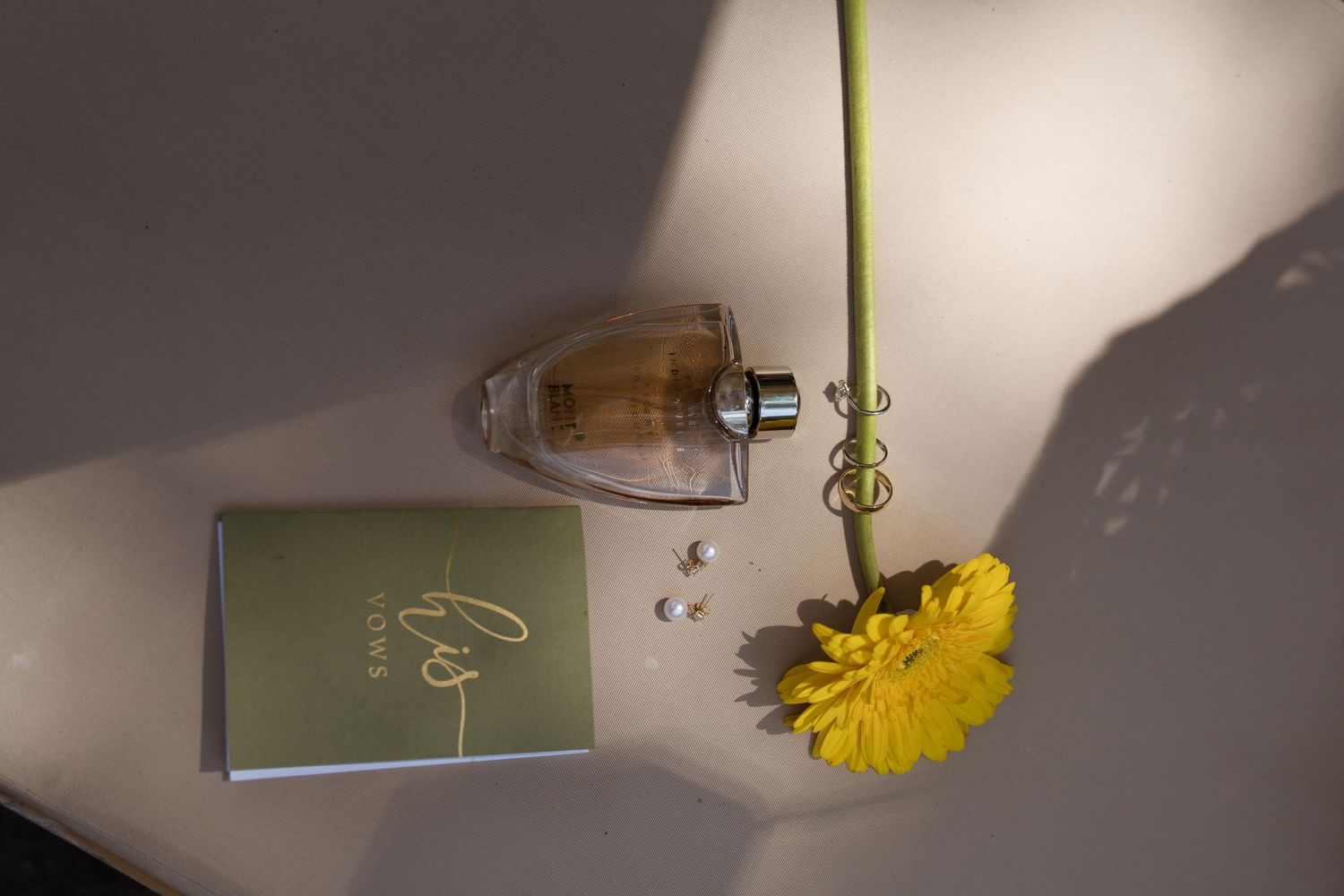 Flat lay of perfume bottle, green card, pearl earrings, and yellow flower on white surface.
