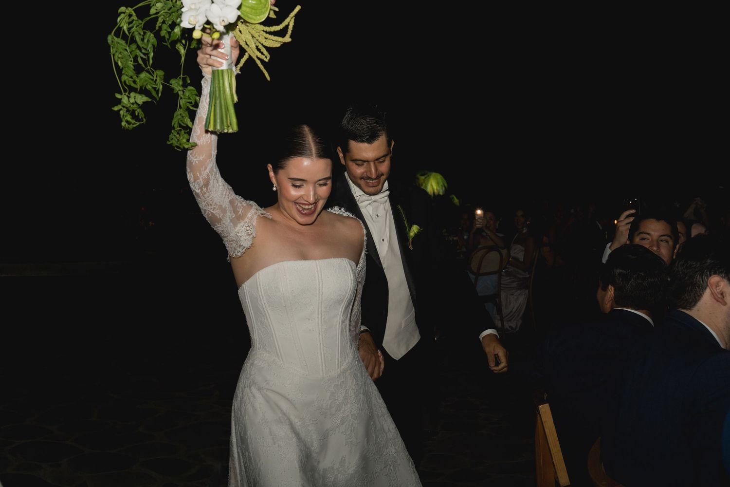 Radiant bride raises her white bouquet triumphantly while walking with groom at their wedding reception.