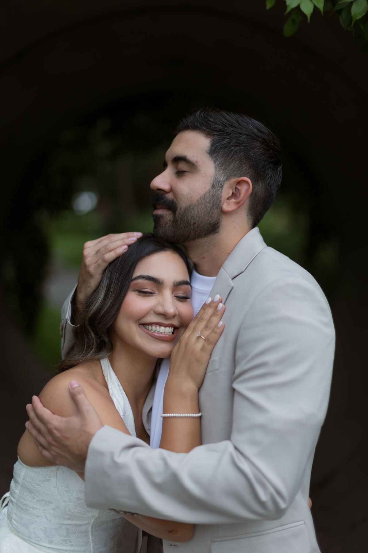 Groom in beige suit tenderly embraces smiling bride under dark leafy archway outdoors.