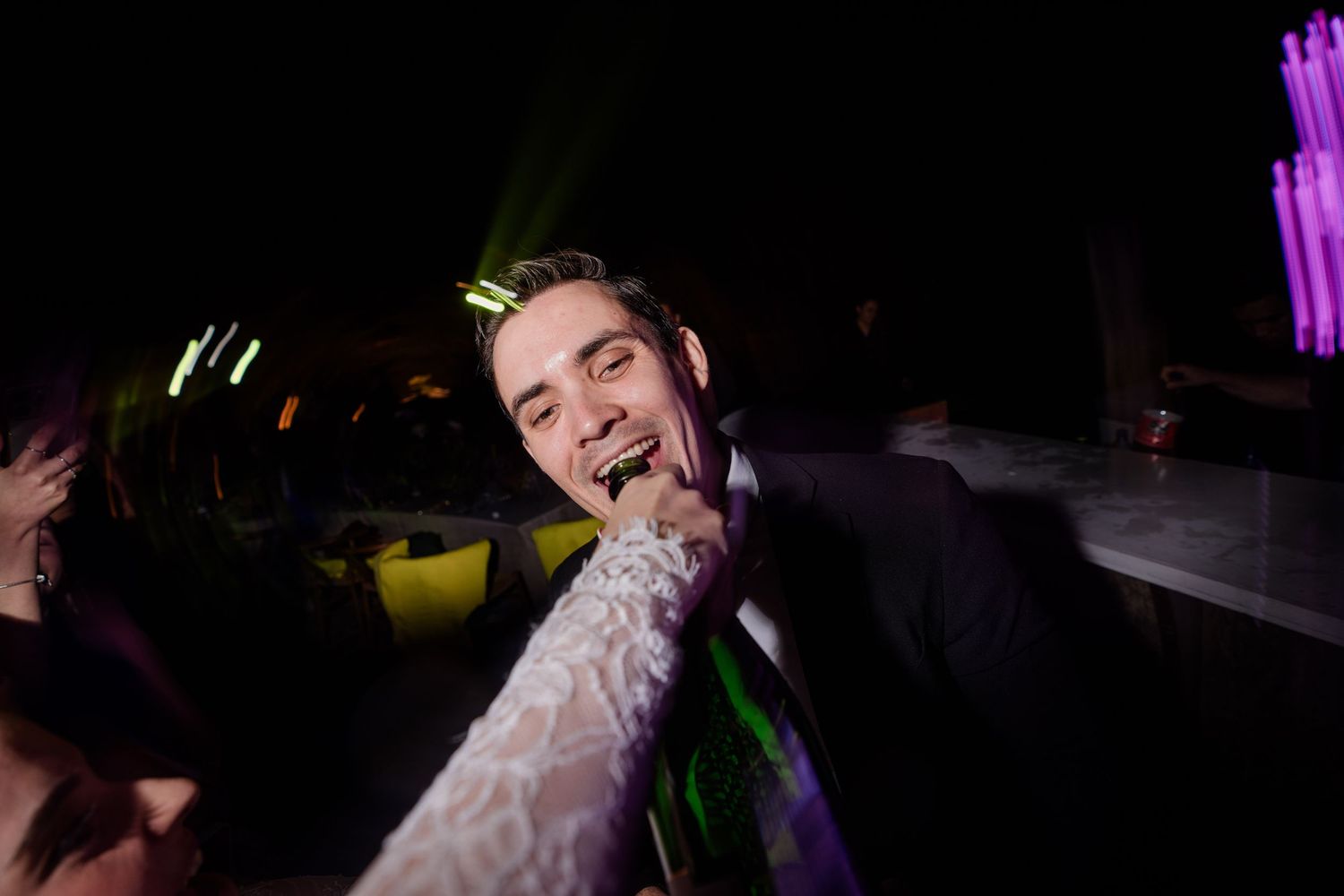 Groom in black suit dancing joyfully under purple and green lights at wedding reception.