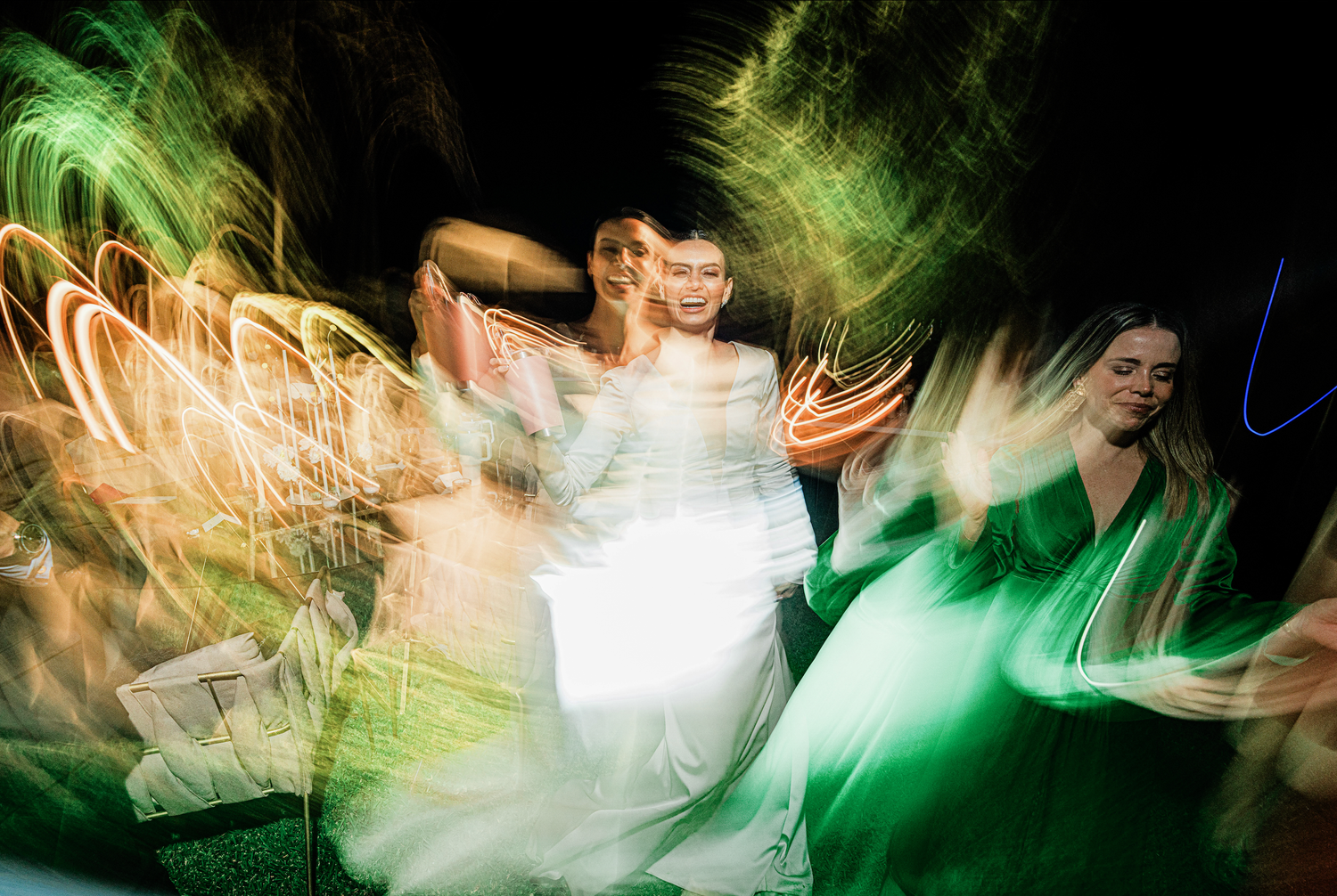 Bride in white gown dances joyfully with guests in green dresses captured with artistic motion blur effect.