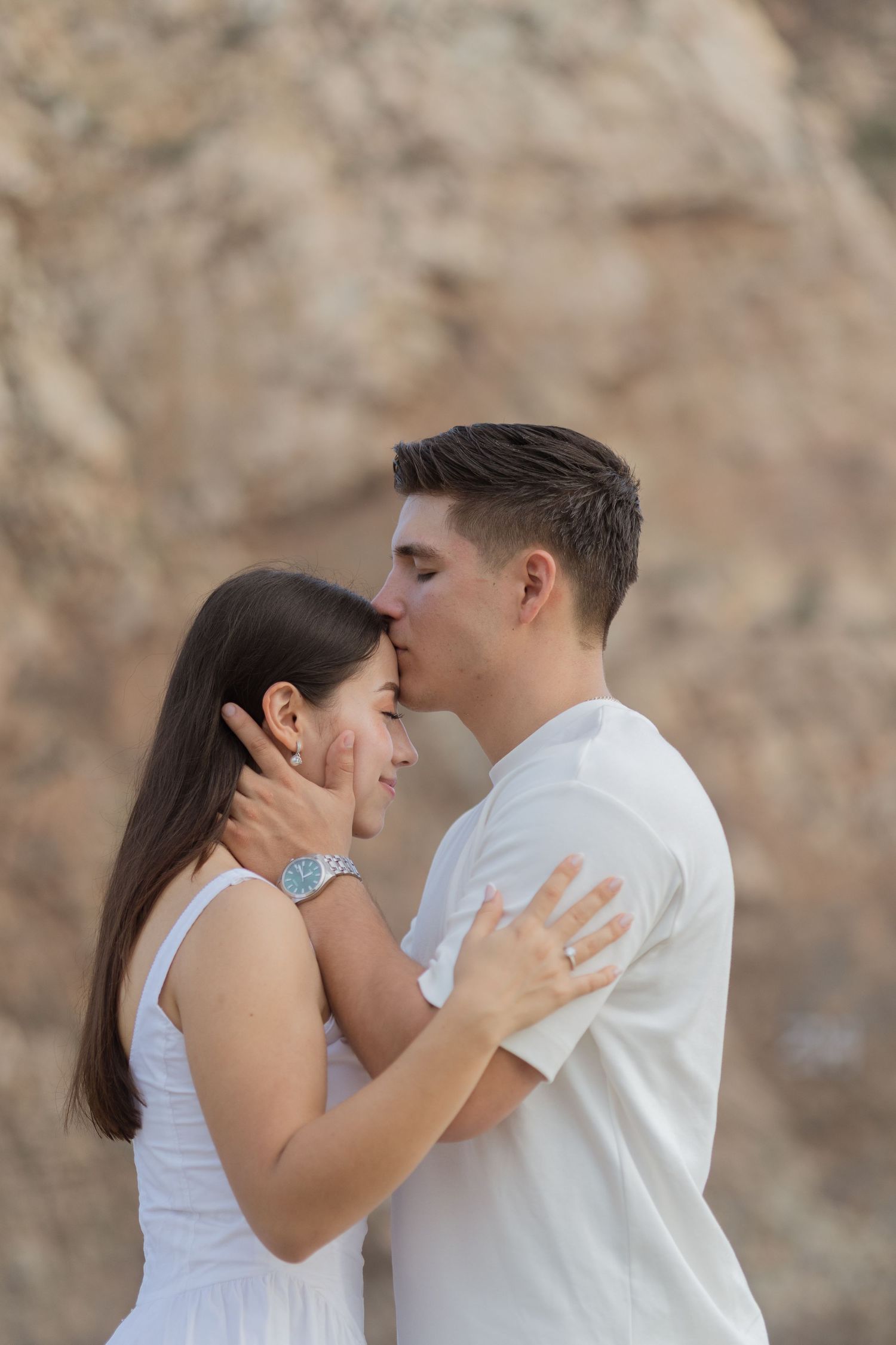 A young couple in white outfits sharing a tender forehead kiss against a rocky background outdoors.