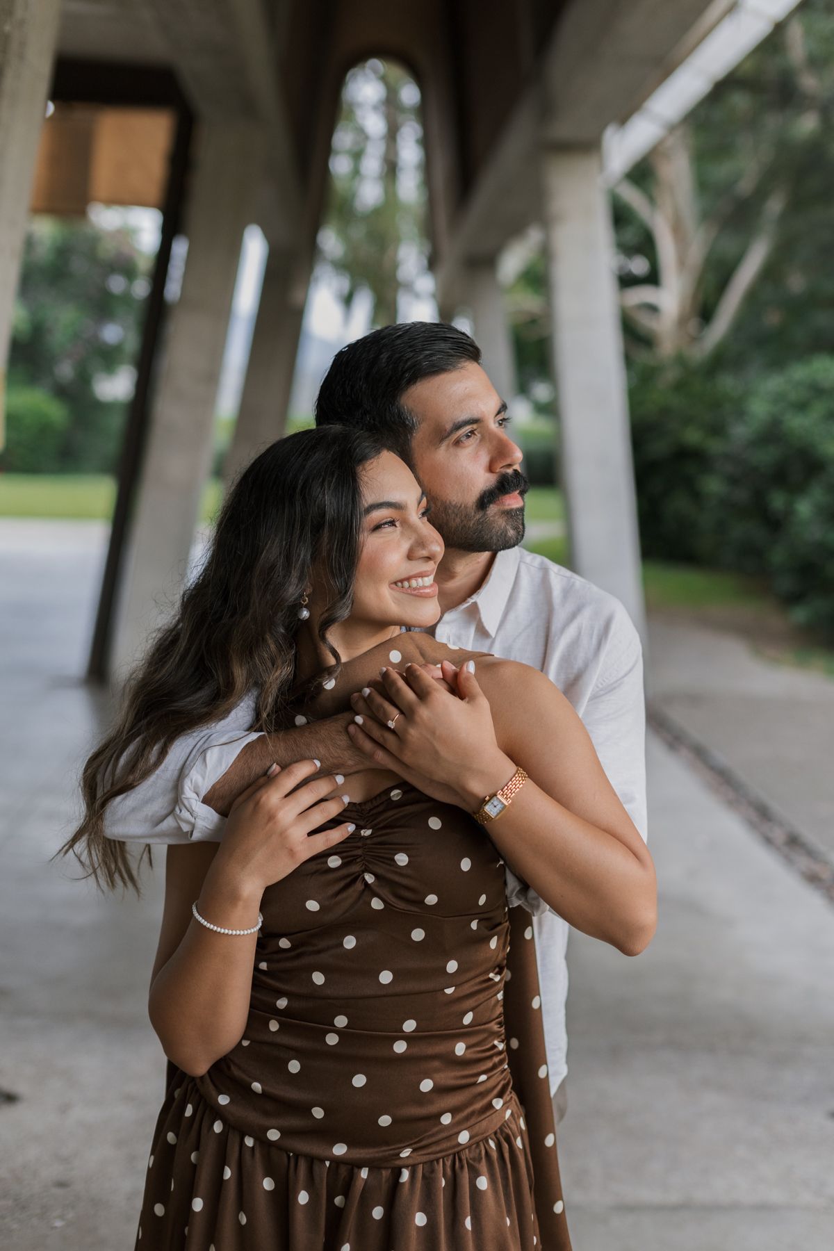 Couple embracing under concrete structure, man in white shirt hugging woman in brown polka dot dress outdoors.