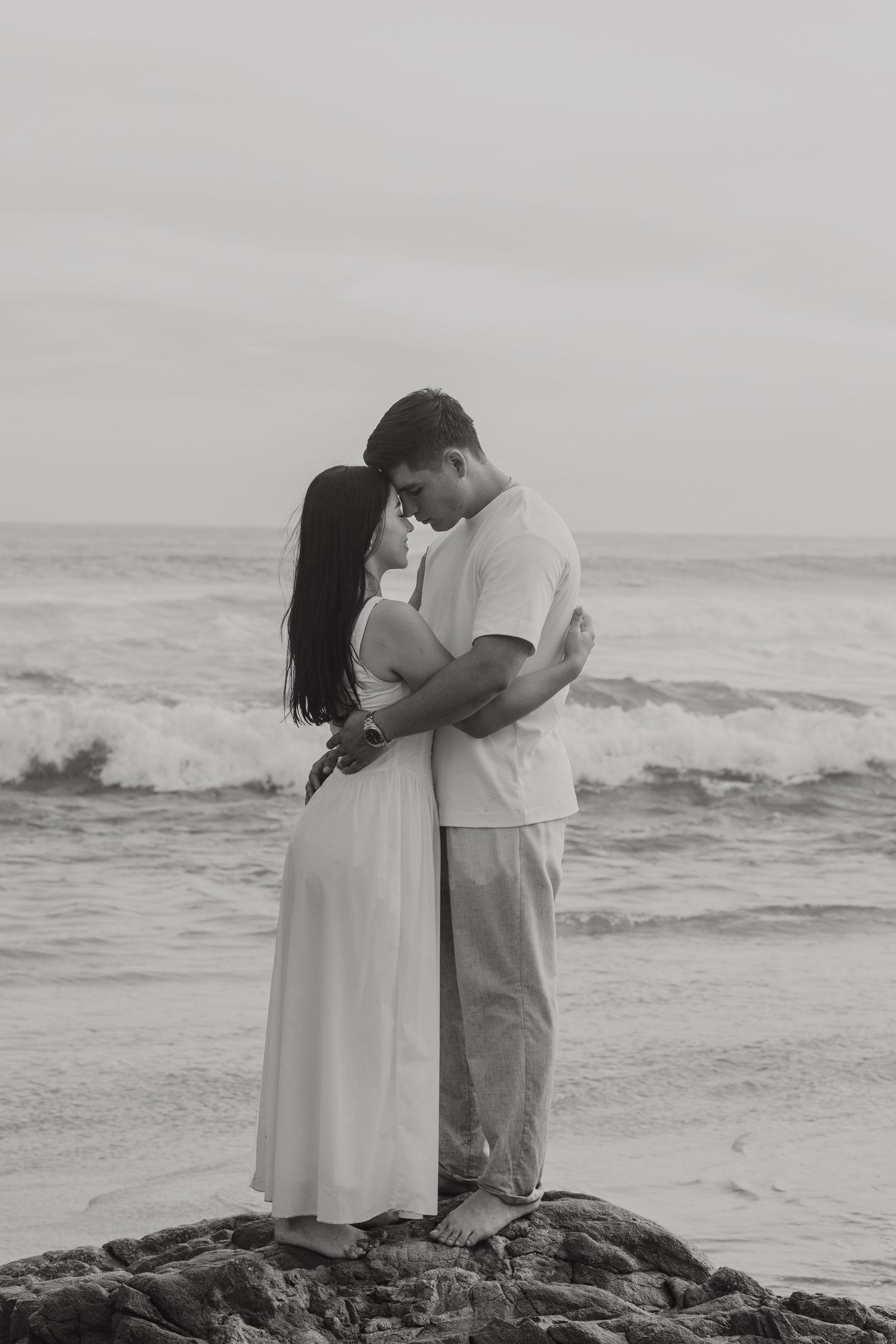 Black and white photo of a couple embracing romantically on rocky beach with ocean waves behind them.