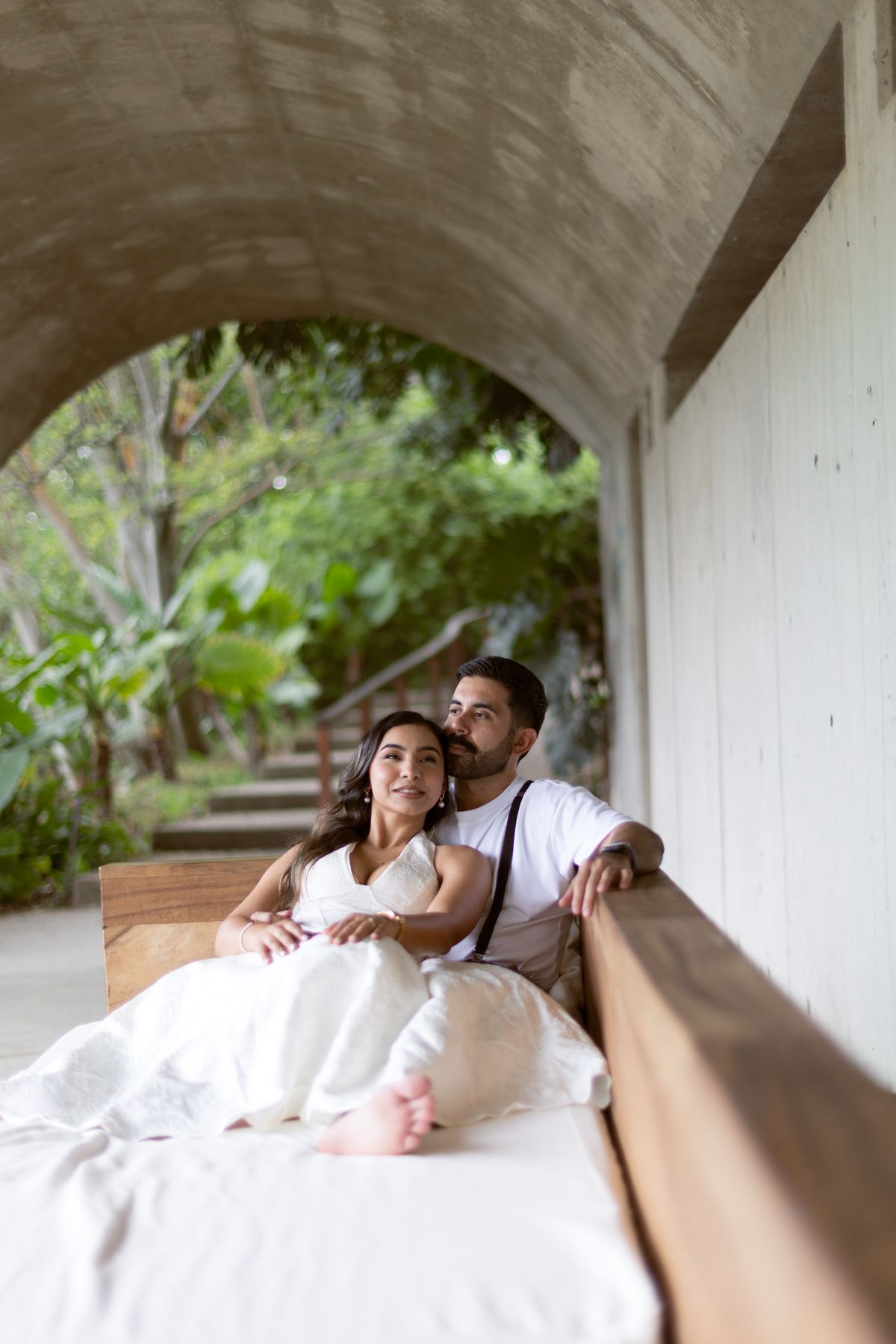Couple relaxing on an outdoor daybed under a concrete arch, surrounded by lush tropical greenery.