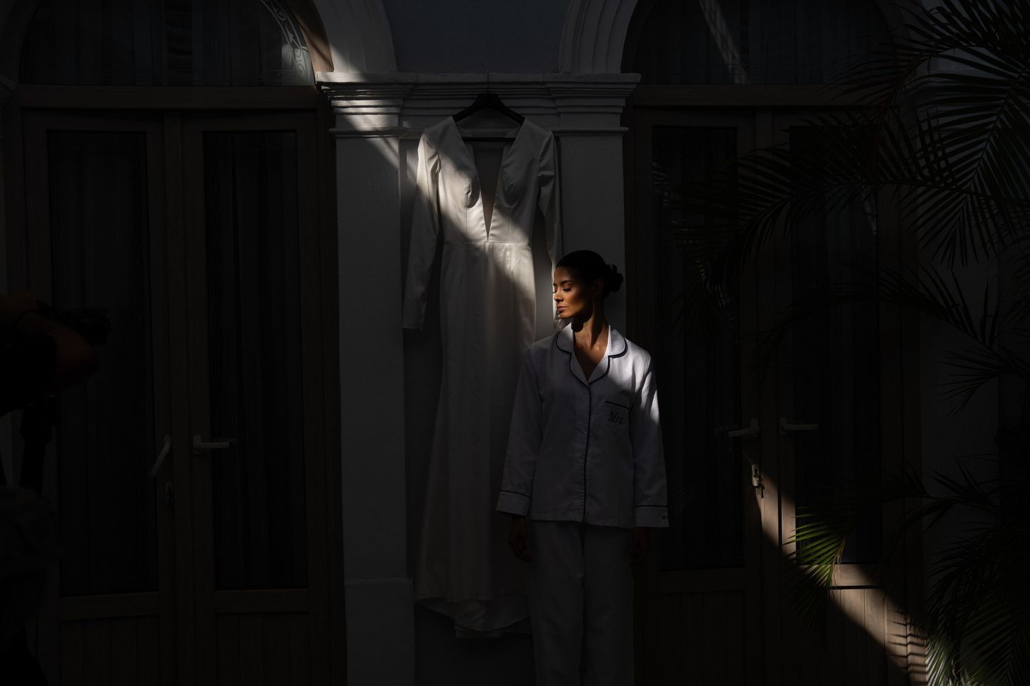 Bride in white suit illuminated by natural light with wedding dress hanging nearby.