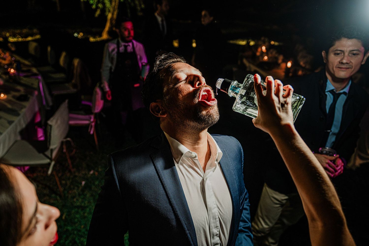 Wedding guest tilts head back drinking from bottle at fun outdoor evening wedding reception celebration.