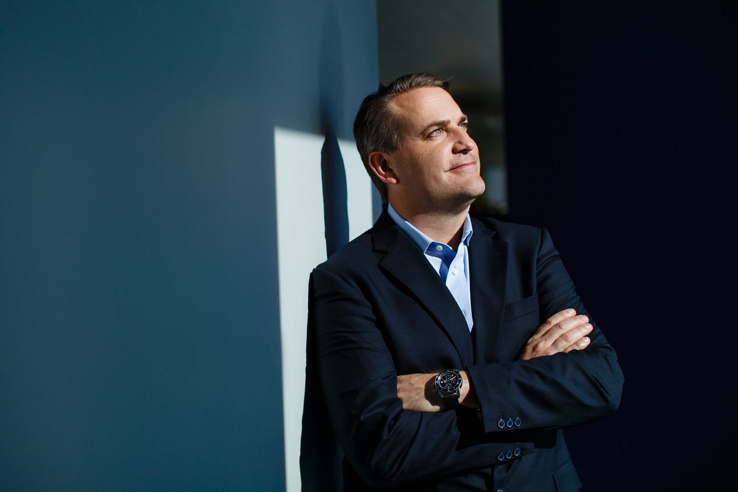 Professional businessman in dark blazer stands with arms crossed, leaning against a blue wall, looking upward confidently.