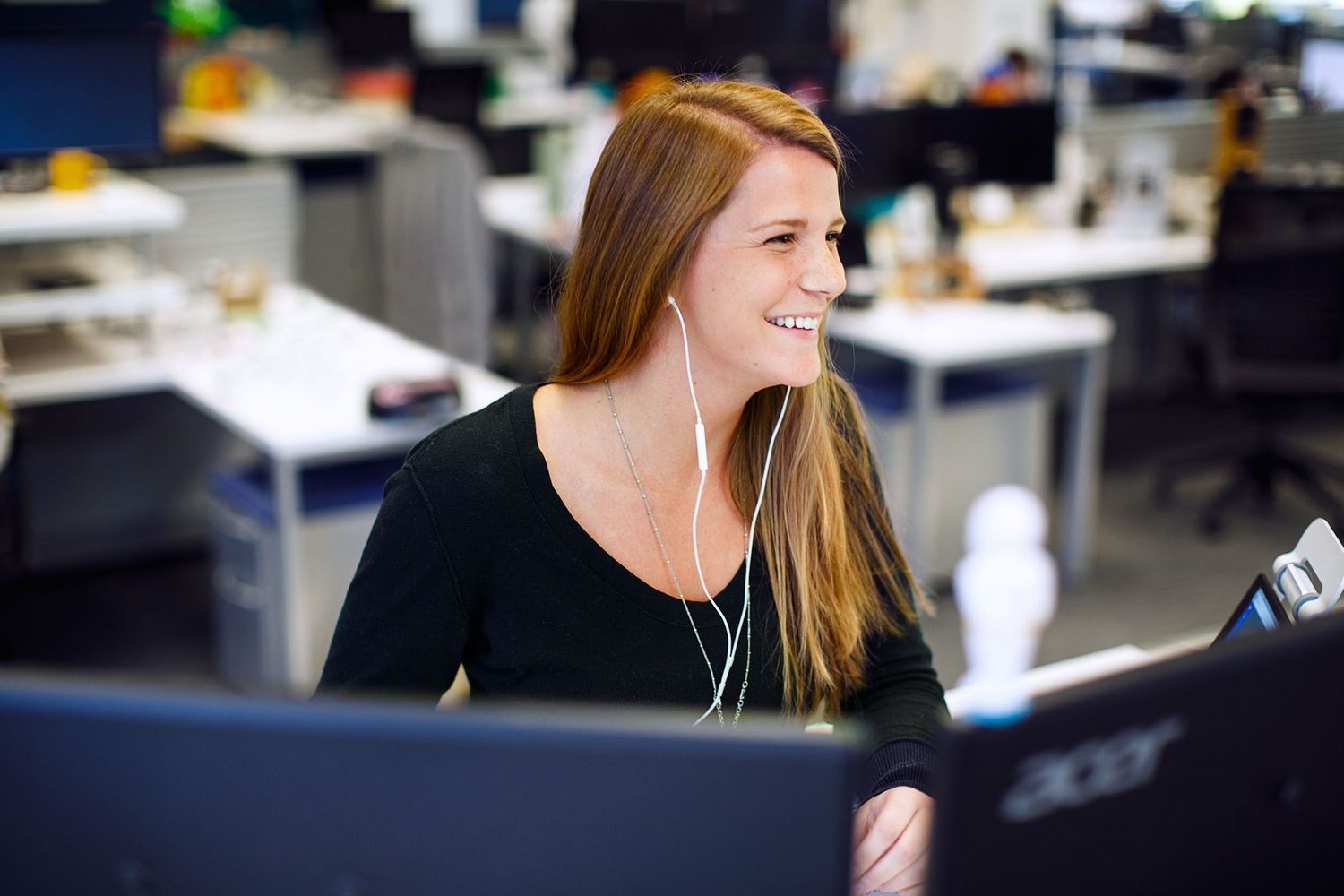 Smiling woman with red hair wearing earphones working at a computer in a modern open-plan office.