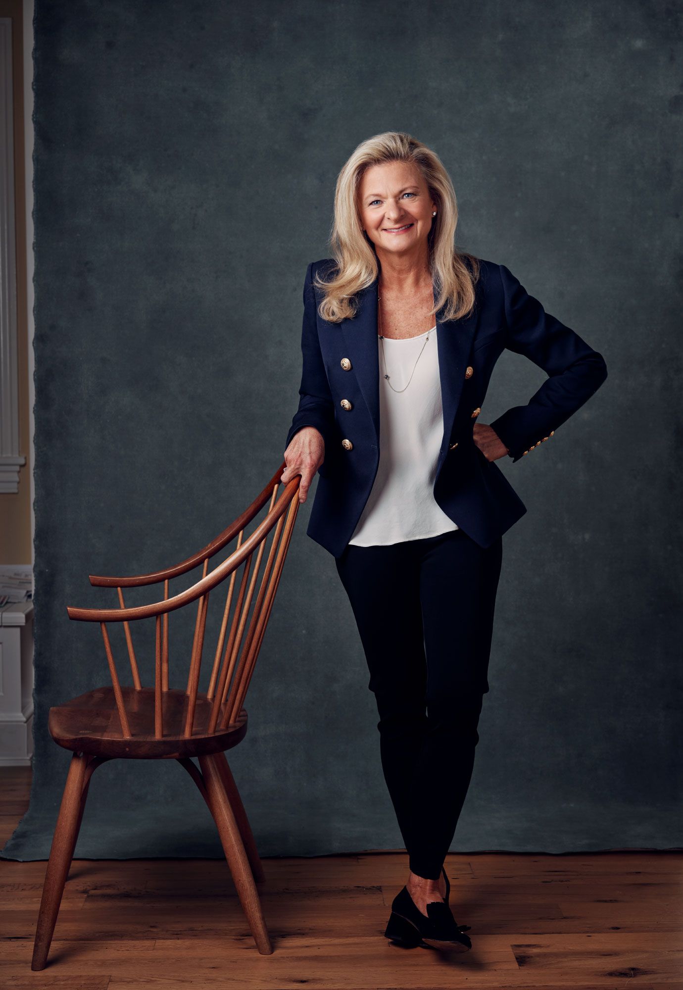 Blonde woman in navy blazer poses beside a wooden Windsor chair on hardwood floors.