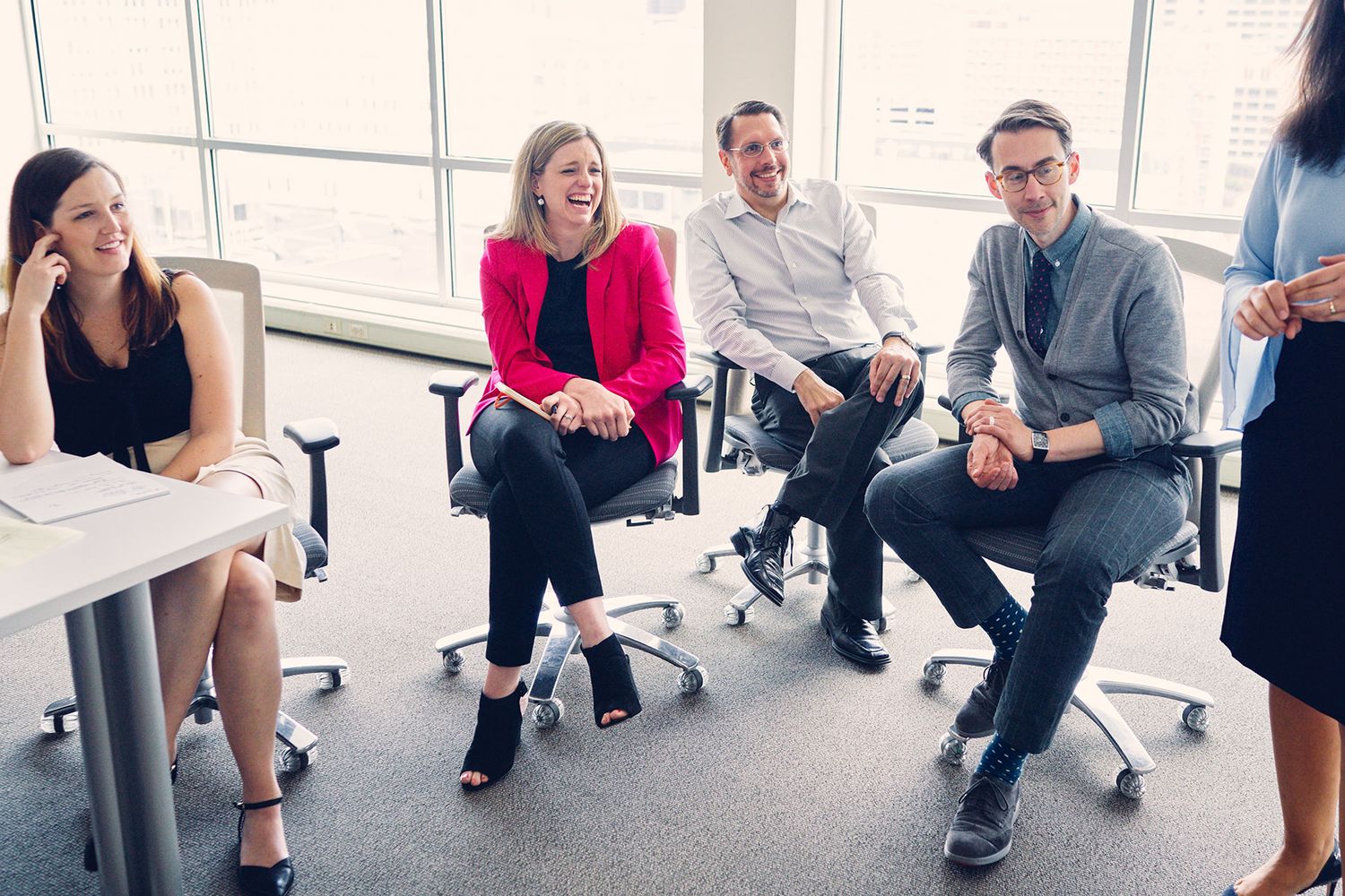 Four smiling business professionals sitting in office chairs having a casual meeting by bright windows.