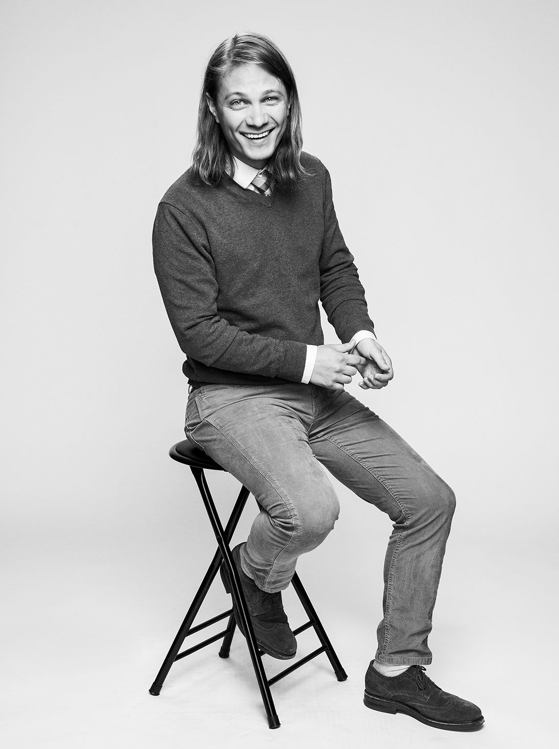 Black and white portrait of a young man with long hair sitting on a stool laughing.