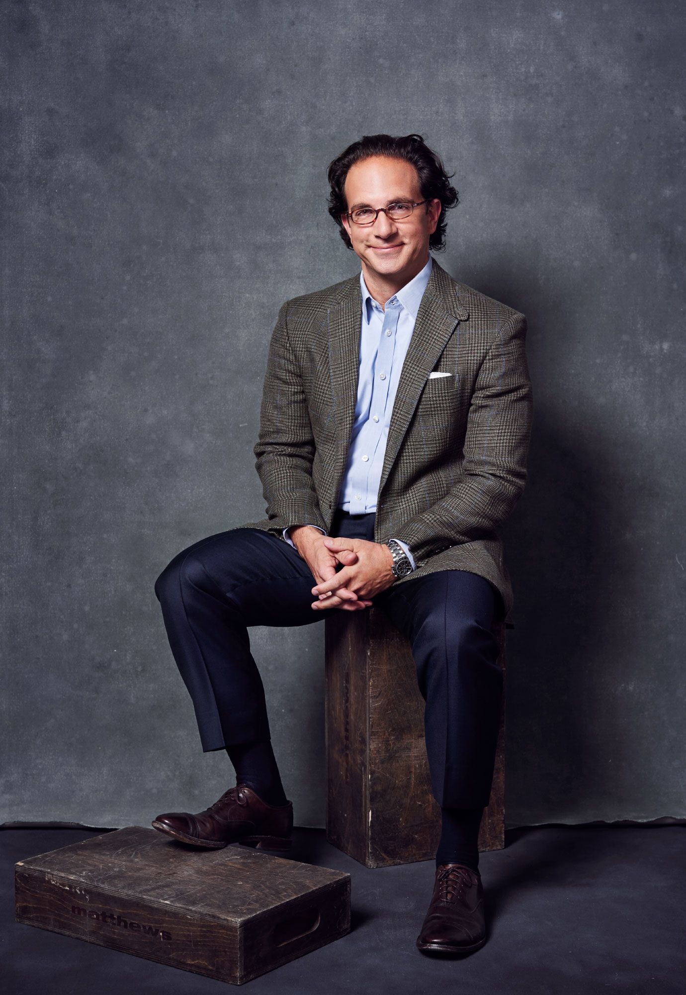 Professional man in tweed blazer sits on wooden box for polished business portrait on gray background.