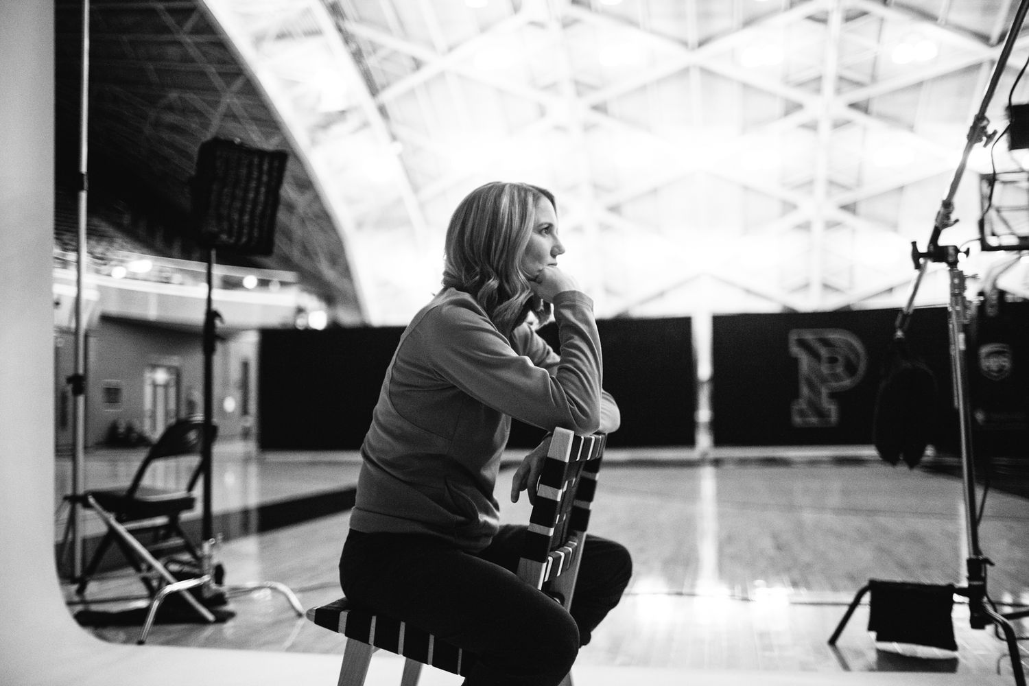 Black and white photo of a woman sitting on a stool in a gymnasium, looking thoughtfully to the side.